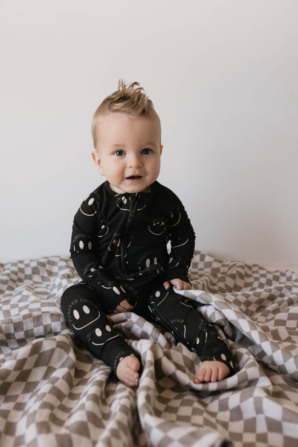 A baby with a mohawk hairstyle sits on a checkered blanket, dressed in Forever French Baby's Bamboo Zip Pajamas in Charcoal & White ff Smile, which are hypo-allergenic. The baby's outfit features white smiley faces and they gaze towards the camera with a curious expression. The neutral background keeps all attention on the baby.