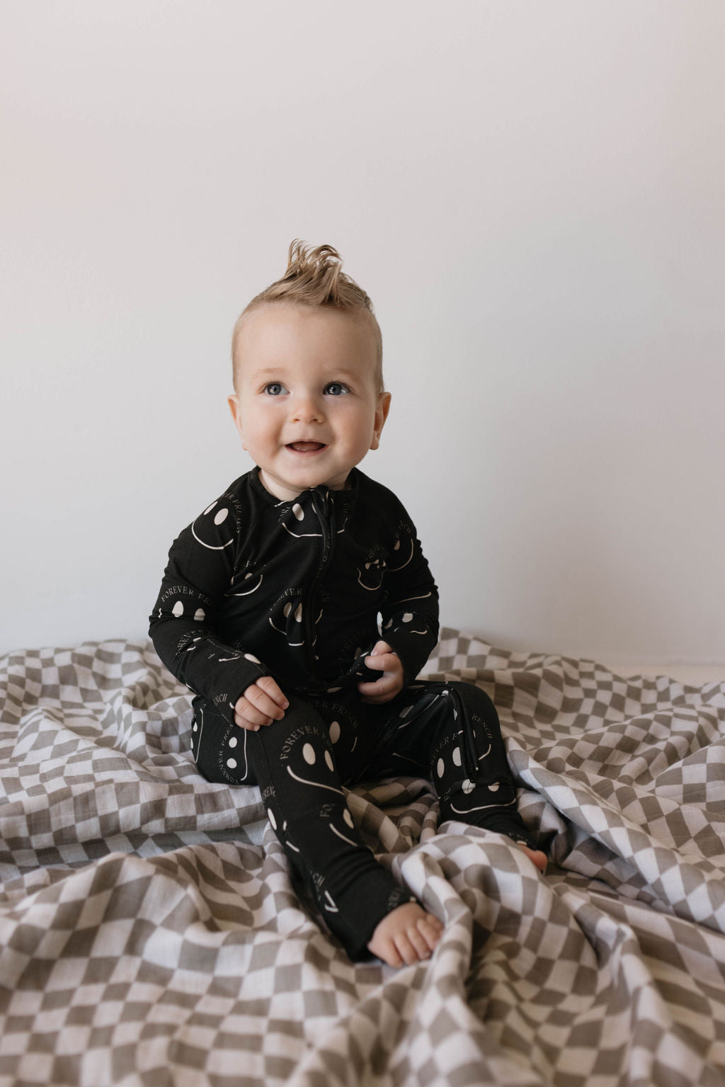 A baby with light hair sits on a gray and white checkered blanket. The baby, dressed in the hypo-allergenic, breathable Bamboo Zip Pajamas in Charcoal & White ff Smile by forever french baby—a black onesie adorned with white abstract shapes—smiles while looking ahead. The background is plain white.