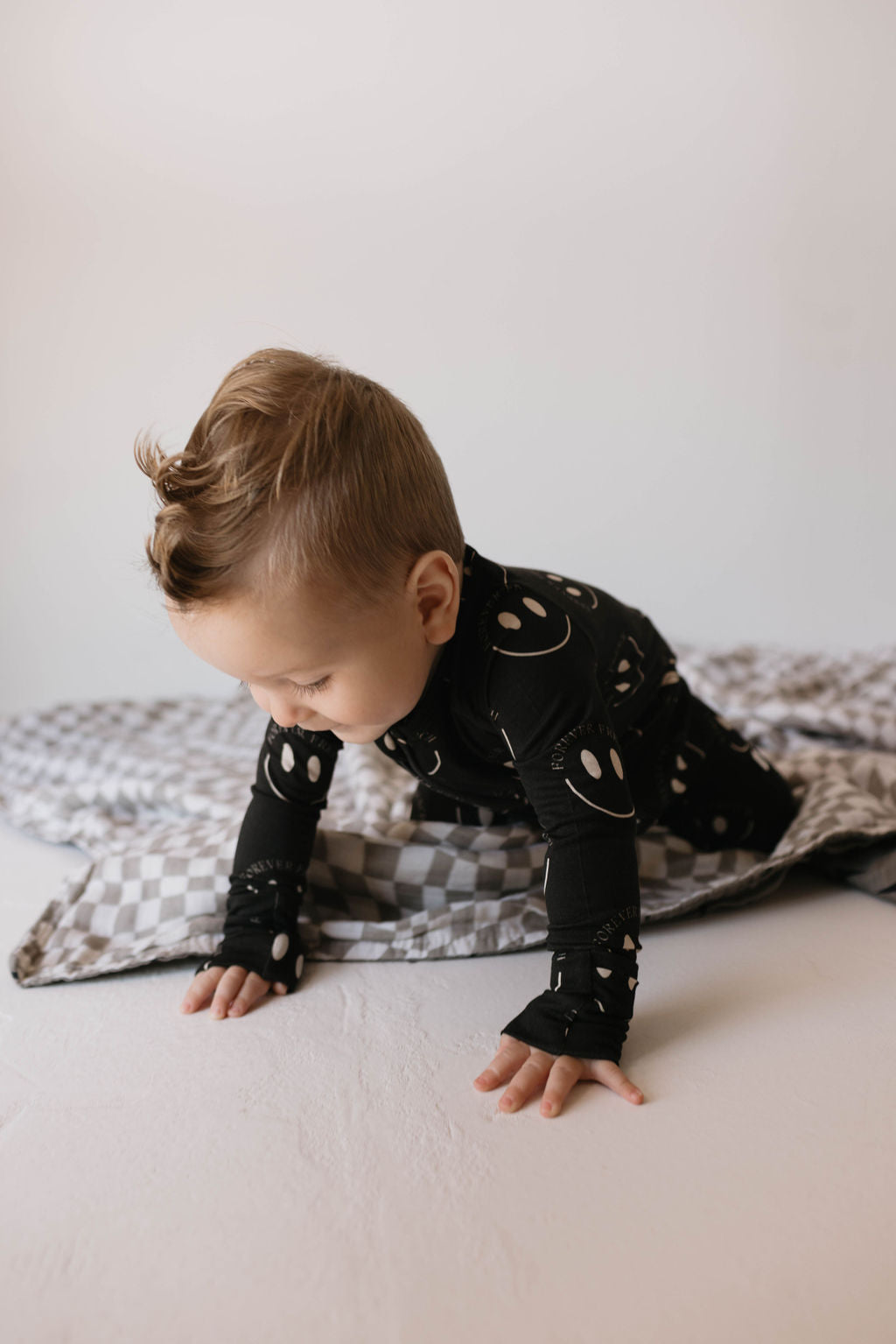 A small child with short light brown hair is crawling on a white surface. The child is wearing black Bamboo Zip Pajamas | Charcoal & White 'ff Smile' from forever french baby, decorated with white smiley faces. Underneath them is a gray and white checkered blanket. The background is plain and white.