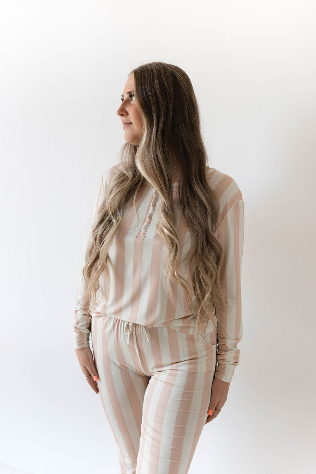 A person stands against a plain white background, wearing the Women's Bamboo Pajamas | Candy Stripe by forever french baby. Crafted from breathable bamboo fabric, this set includes a long-sleeved, button-up top and matching pants adorned with a charming light pink and white vertical Candy Stripe pattern. They have long, wavy hair and are looking to the side.