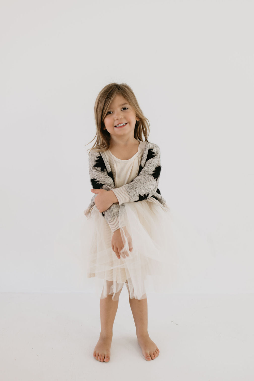 A young girl stands barefoot against a plain white background, wearing a white dress with a fluffy tulle skirt and Forever French Baby's Cozy Cardigan in the Silent Night design. Her arms are crossed, and she has a cheerful expression.