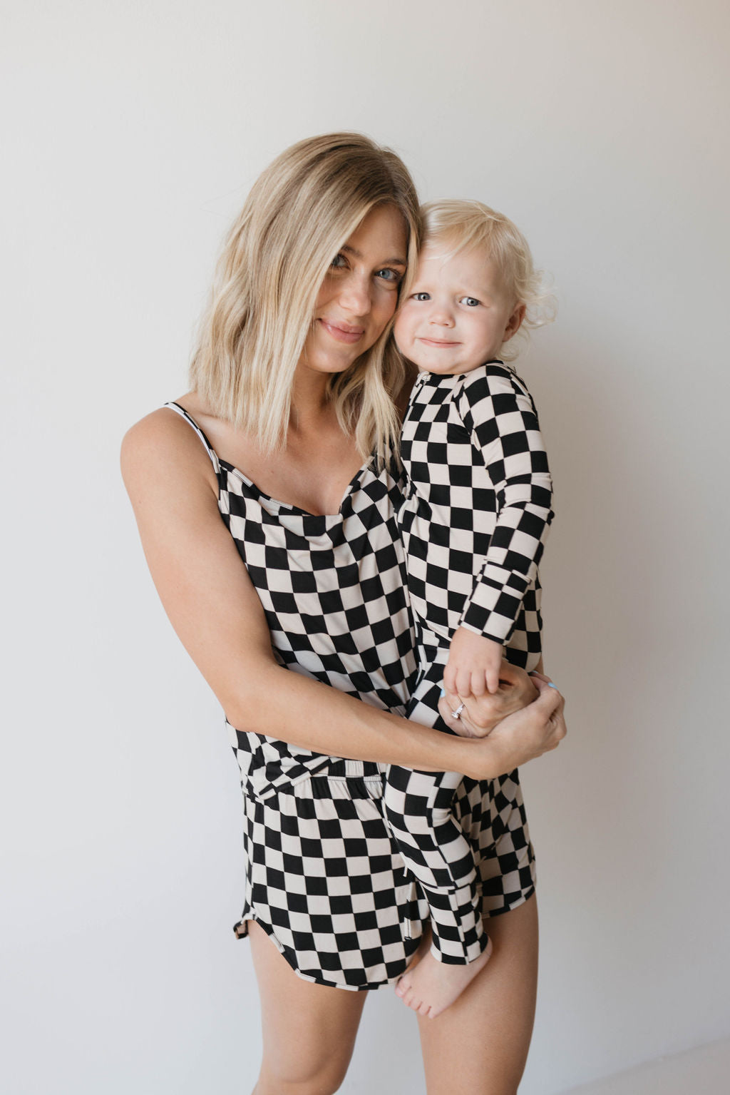 A blonde woman, dressed in a black and white checkered romper, holds a young blonde child clad in matching "Bamboo Zip Pajamas | Black Checkerboard" from forever french baby. Both are smiling against a plain white background.