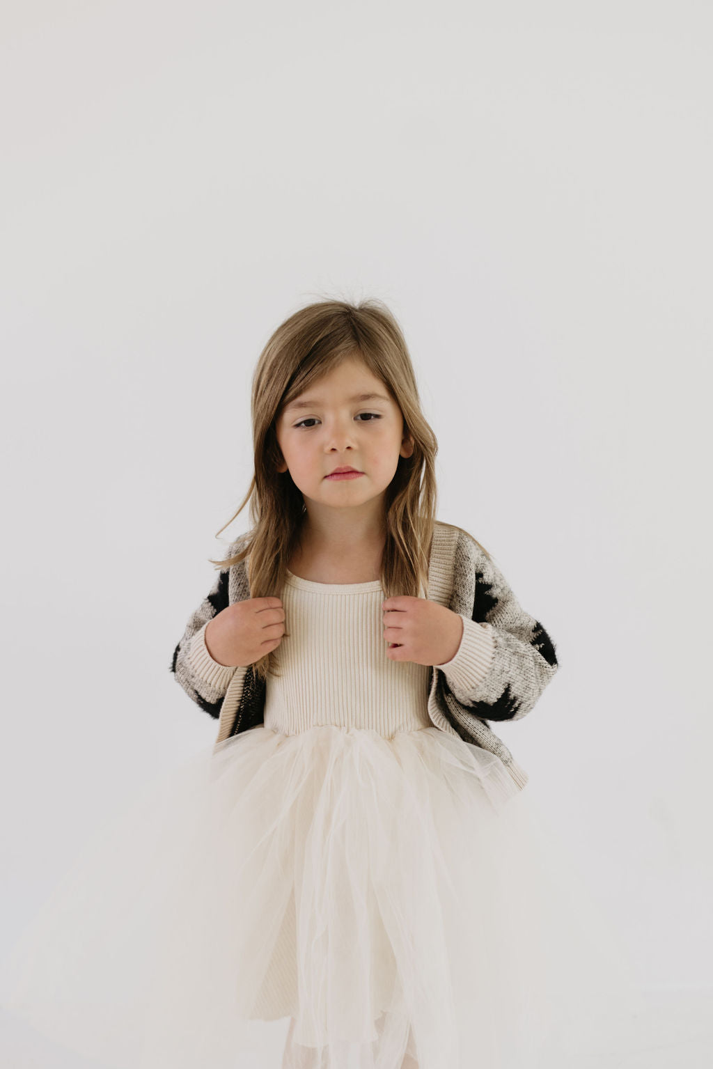 A young girl stands against a plain white background, wearing a white dress with a light, textured skirt and the Cozy Cardigan in Silent Night from forever french baby. Her long hair is loose, and she gazes directly at the camera with a neutral expression.