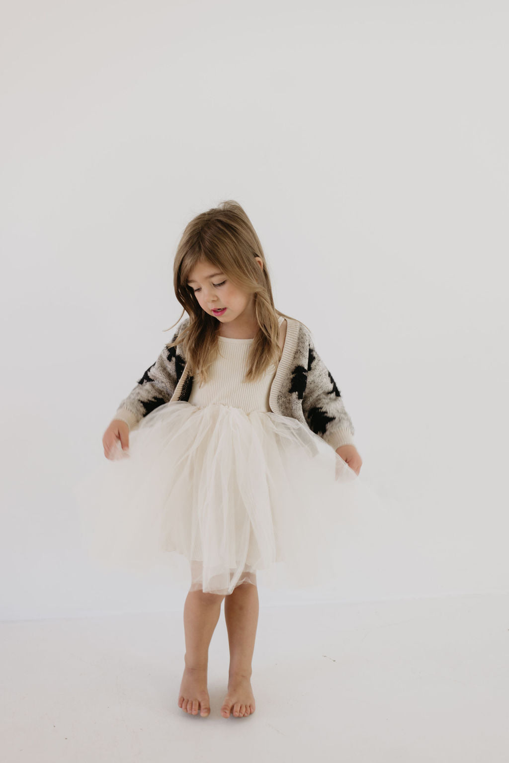 A young girl stands barefoot against a plain background, wearing a fluffy white dress and the Cozy Cardigan in Silent Night. The softest cardigan drapes gently over her shoulders as she looks down, holding the sides of her dress lightly—an enchanting vision reminiscent of Forever French Baby elegance.