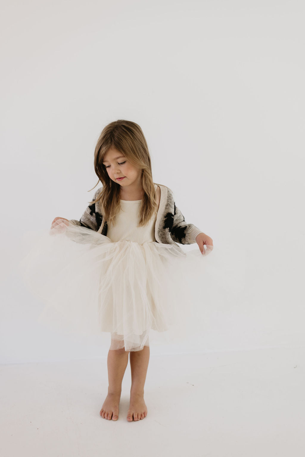 A young girl stands barefoot against a white background, wearing a cream tulle dress and the Cozy Cardigan from Forever French Baby with its subtle Silent Night Christmas tree design. She looks down, gently holding the sides of her dress, embodying the elegance of Forever French Baby style.