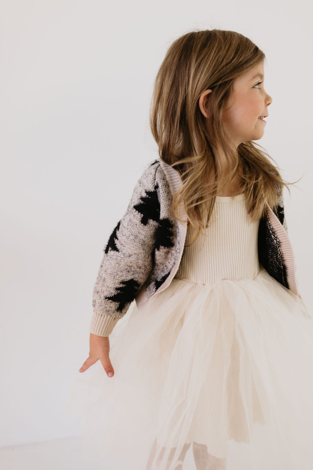 A young girl in a cream-colored tulle dress and a Forever French Baby Cozy Cardigan in the Silent Night design, featuring a Christmas tree pattern, stands against a plain white background. She has long, light brown hair and appears to be smiling slightly as she looks to her right.