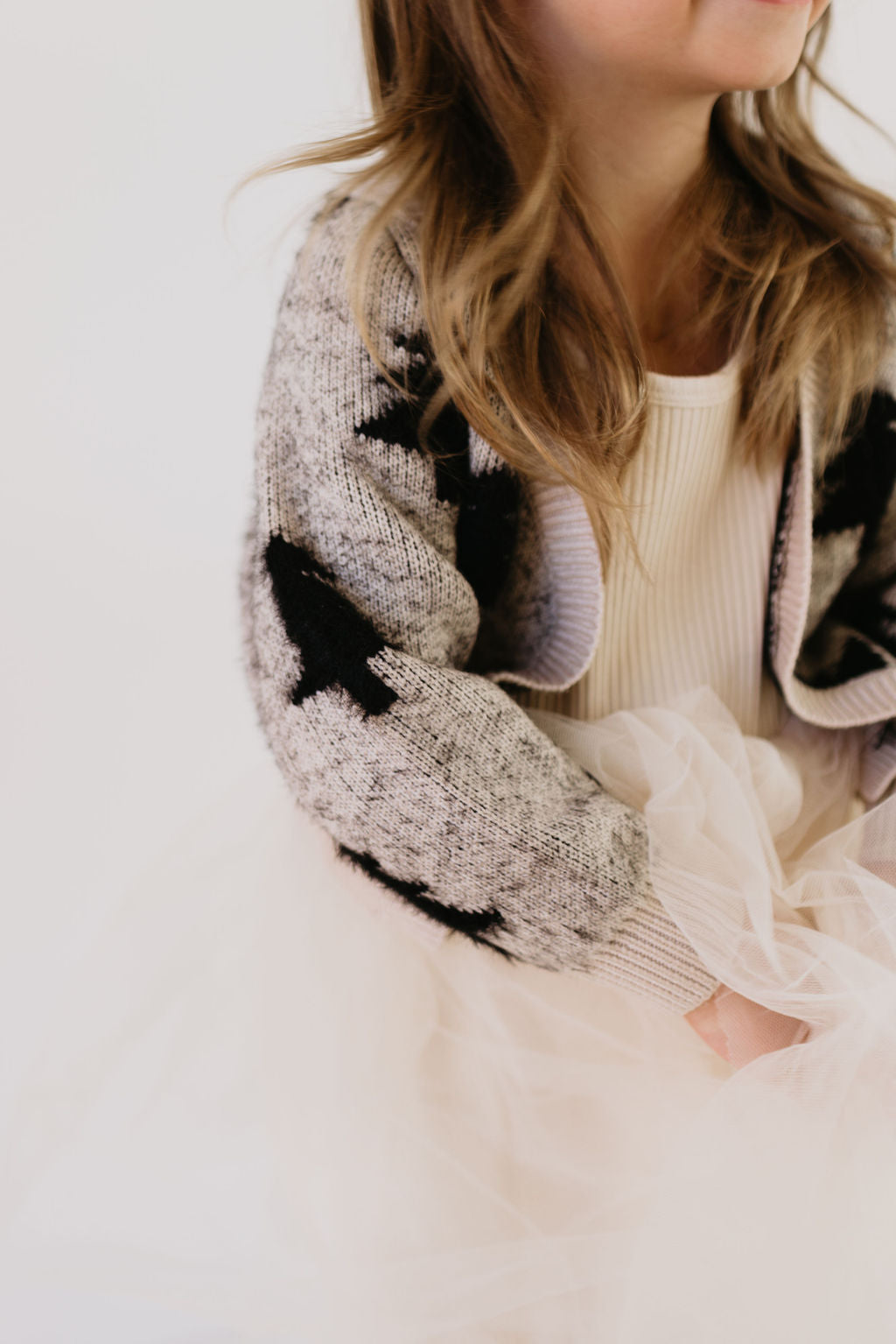 A young girl wearing Forever French Baby's Cozy Cardigan in the Silent Night design smiles slightly, her light brown wavy hair framing her face. She is dressed in a cozy gray sweater adorned with black star patterns and a cream-colored tulle skirt, looking serene against a soft, neutral background.
