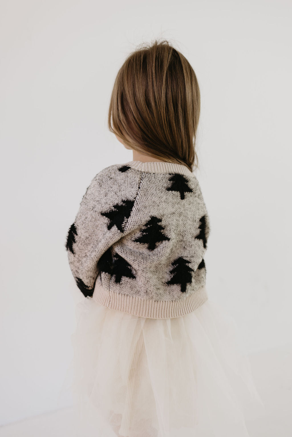 A young child with long hair stands with their back to the camera, wearing the Cozy Cardigan in Silent Night by forever french baby. The cardigan is adorned with a Christmas tree design and paired with a cream-colored tulle skirt, creating an enchanting look set against a plain light background.