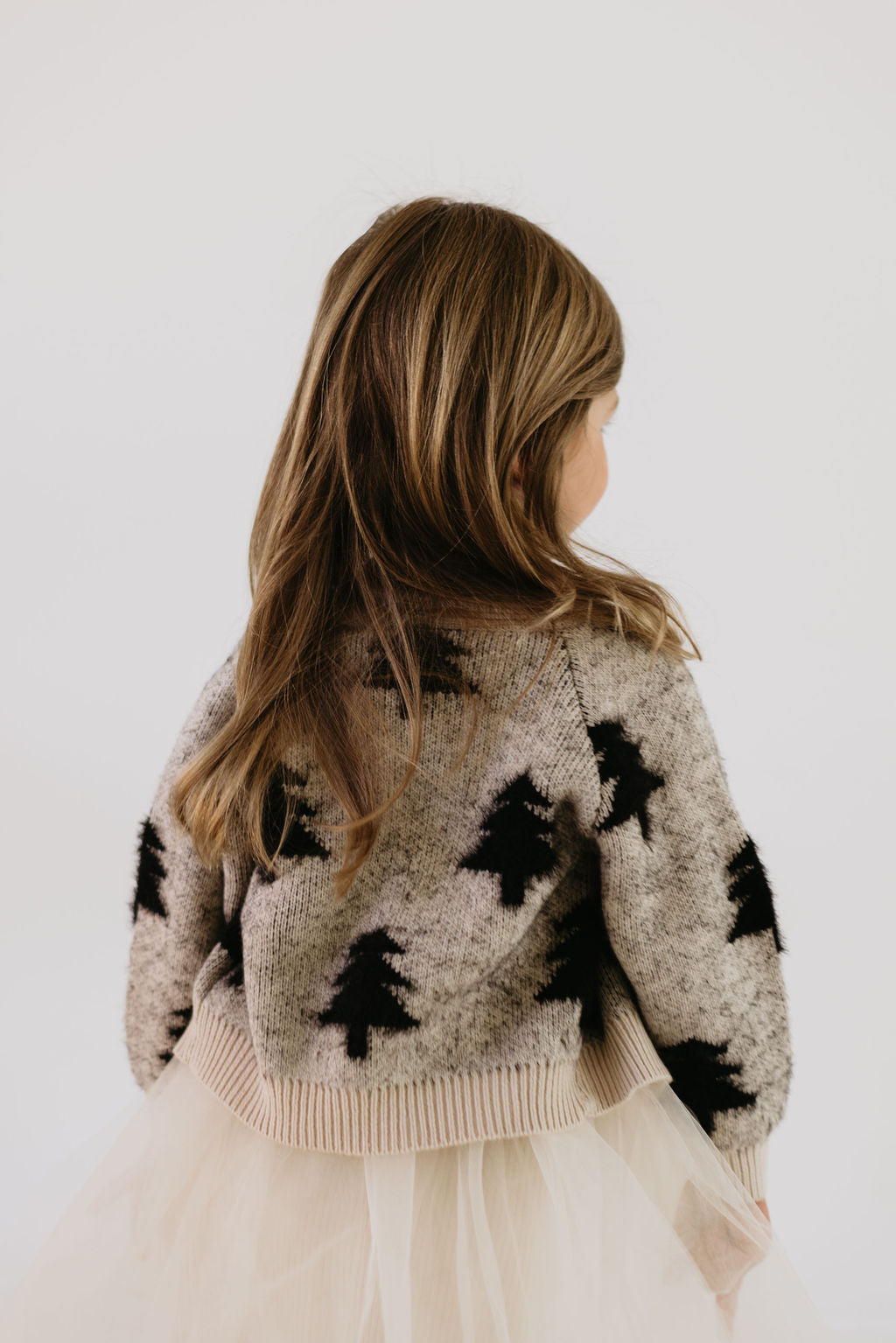 A young girl with long brown hair is wearing the Cozy Cardigan | Silent Night from forever french baby, which is designed with a Christmas tree pattern, and a white tulle skirt. She has her back to the camera against a plain white background.