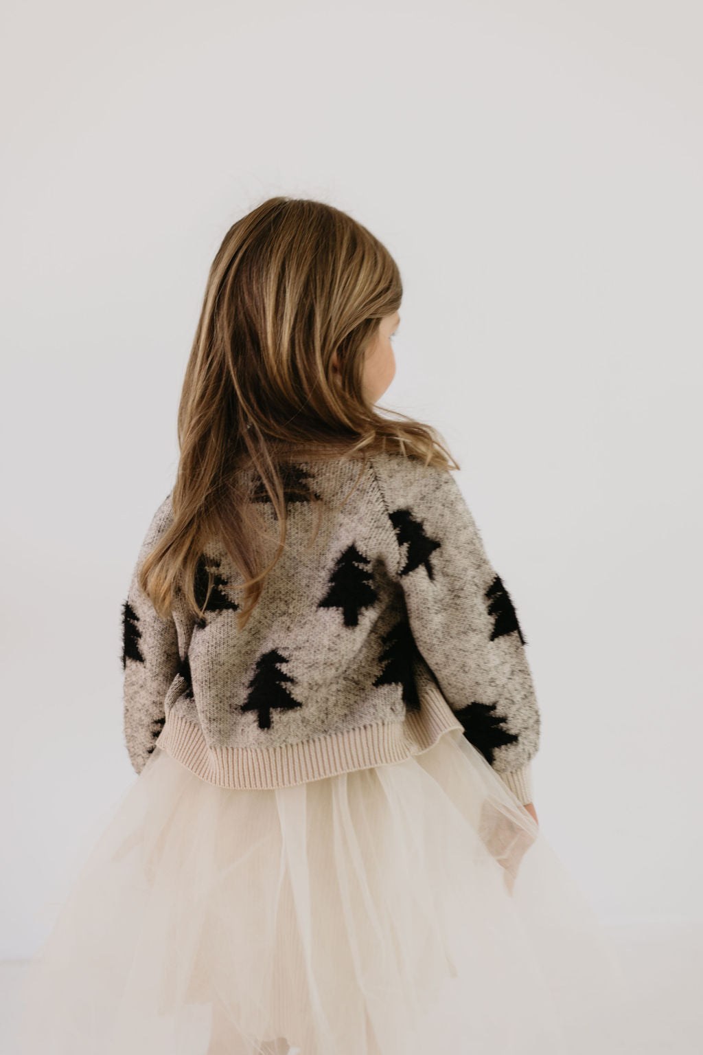A young girl with long brown hair is wearing a Cozy Cardigan from the "Silent Night" collection by forever french baby, paired with a fluffy, cream-colored tulle skirt. She stands against a plain white background, facing away.