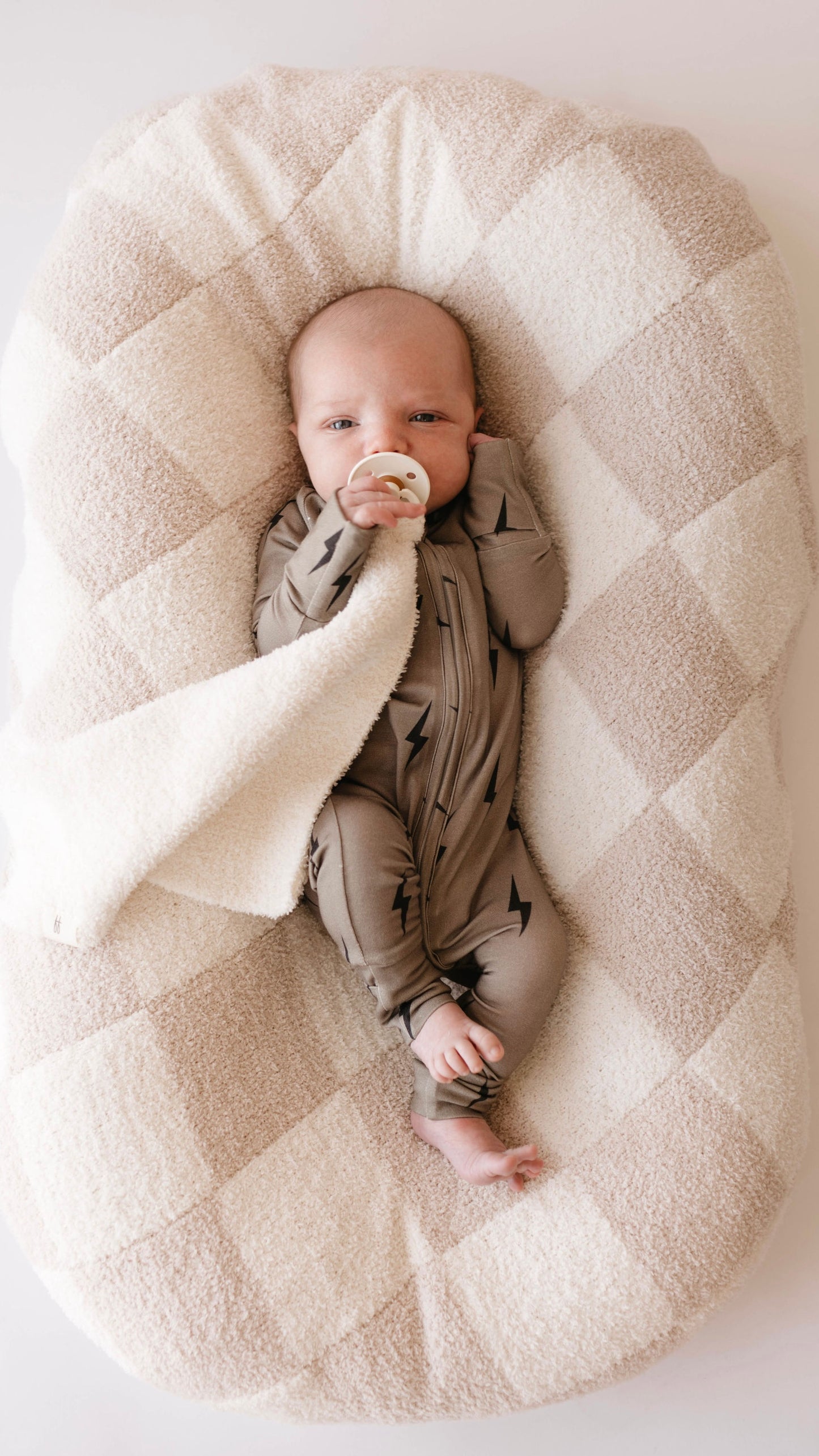 A baby dressed in forever french baby's Bamboo Zip Pajamas in a brown and black lightning bolt pattern lies on a plush, checkered cushion. The baby's left hand rests on their face while the right hand holds a pacifier. A white, hypo-allergenic blanket partially covers the baby.