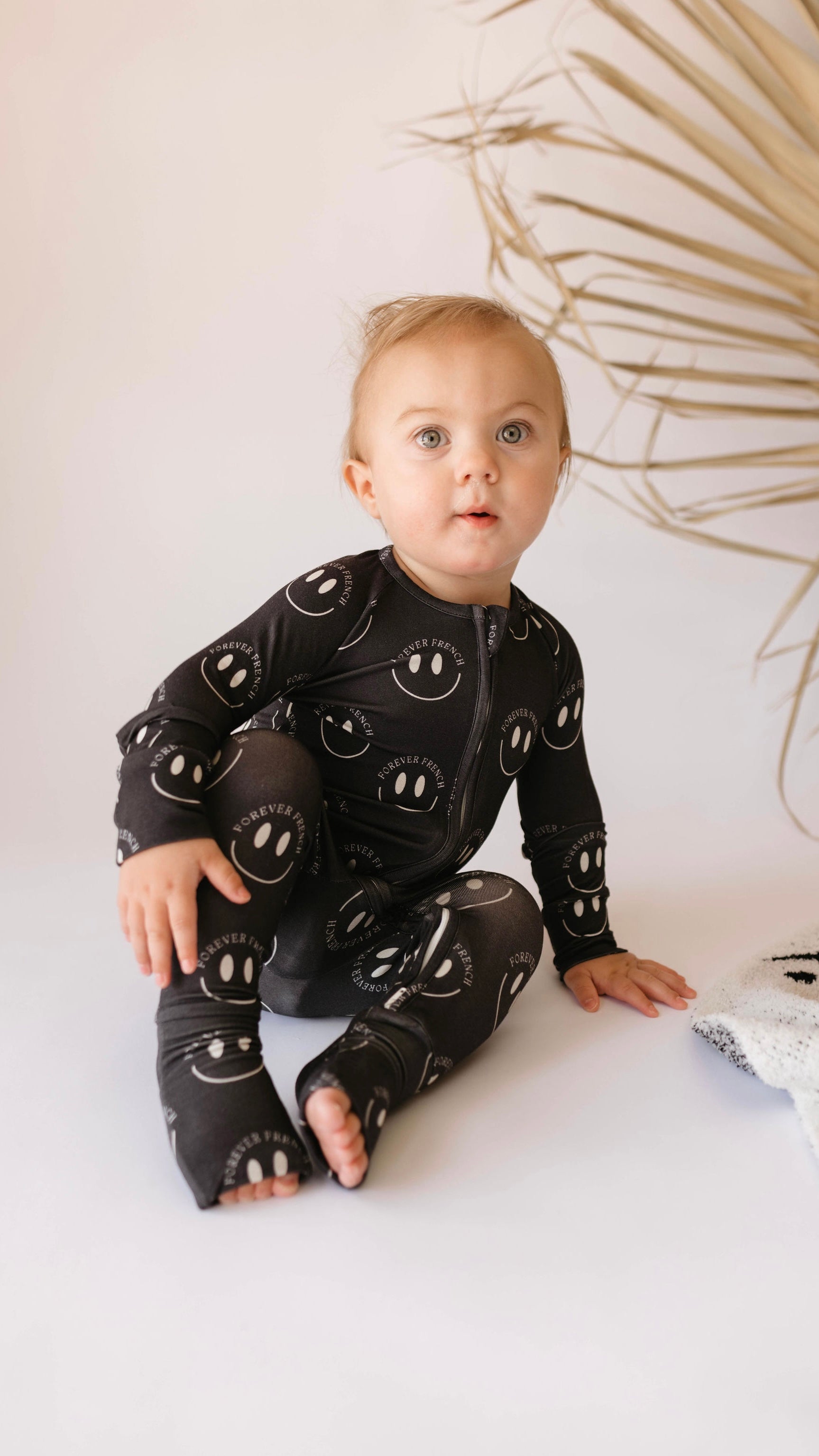 A baby with light hair sits on a white surface, wearing hypoallergenic Bamboo Zip Pajamas by forever french baby, featuring the Charcoal & White ff Smile design. The baby gazes upward with a curious expression. In the background, there is a dried palm leaf. A blanket with a similar smiley face pattern is partially visible.