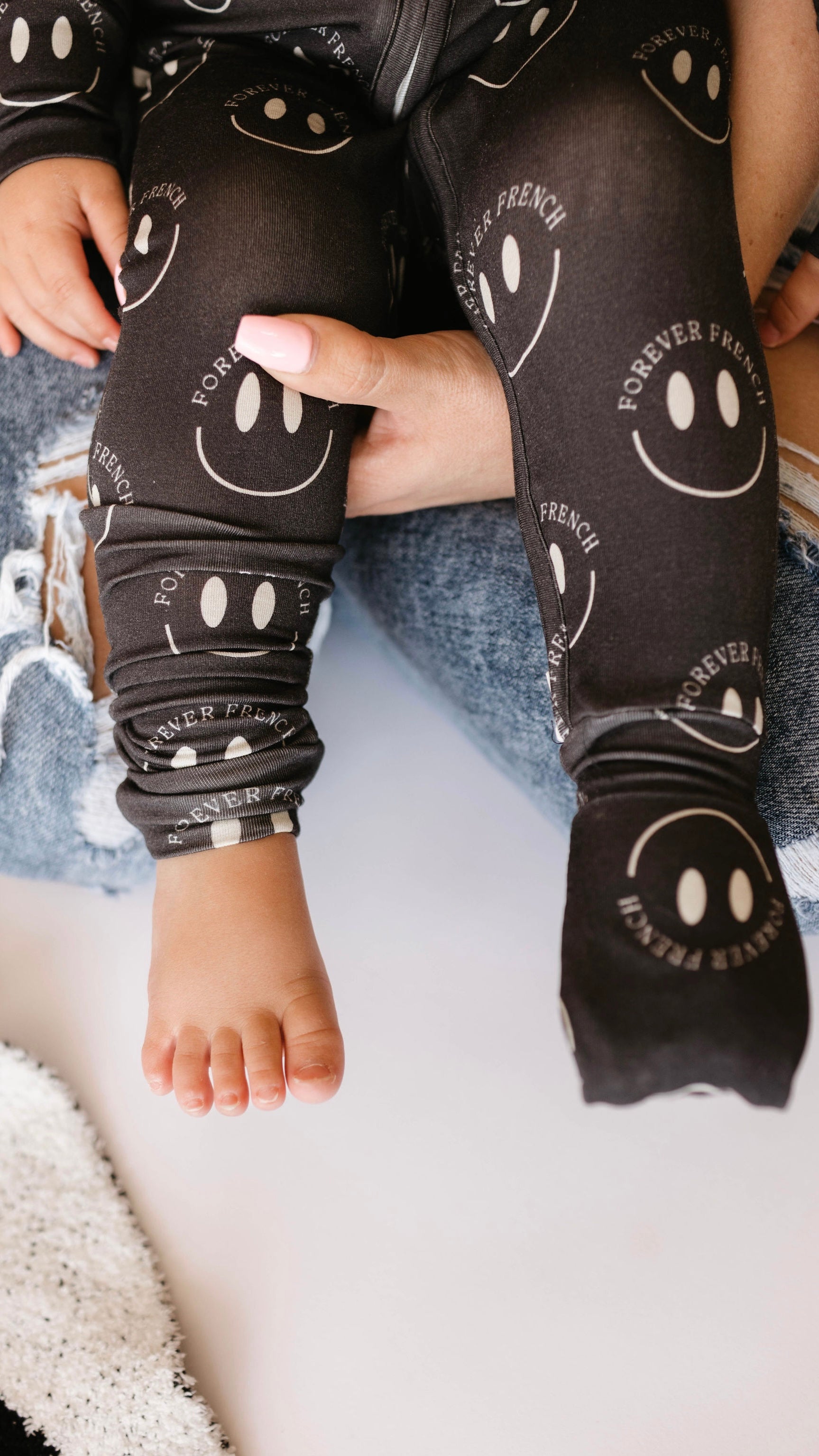 A close-up image of a baby sitting on someone's lap. The baby is wearing Bamboo Zip Pajamas in Charcoal & White ff Smile from the brand forever french baby. The baby's bare foot is visible, resting on the person's jeans, which have a ripped knee. This adorable scene highlights breathable clothing perfect for delicate skin.