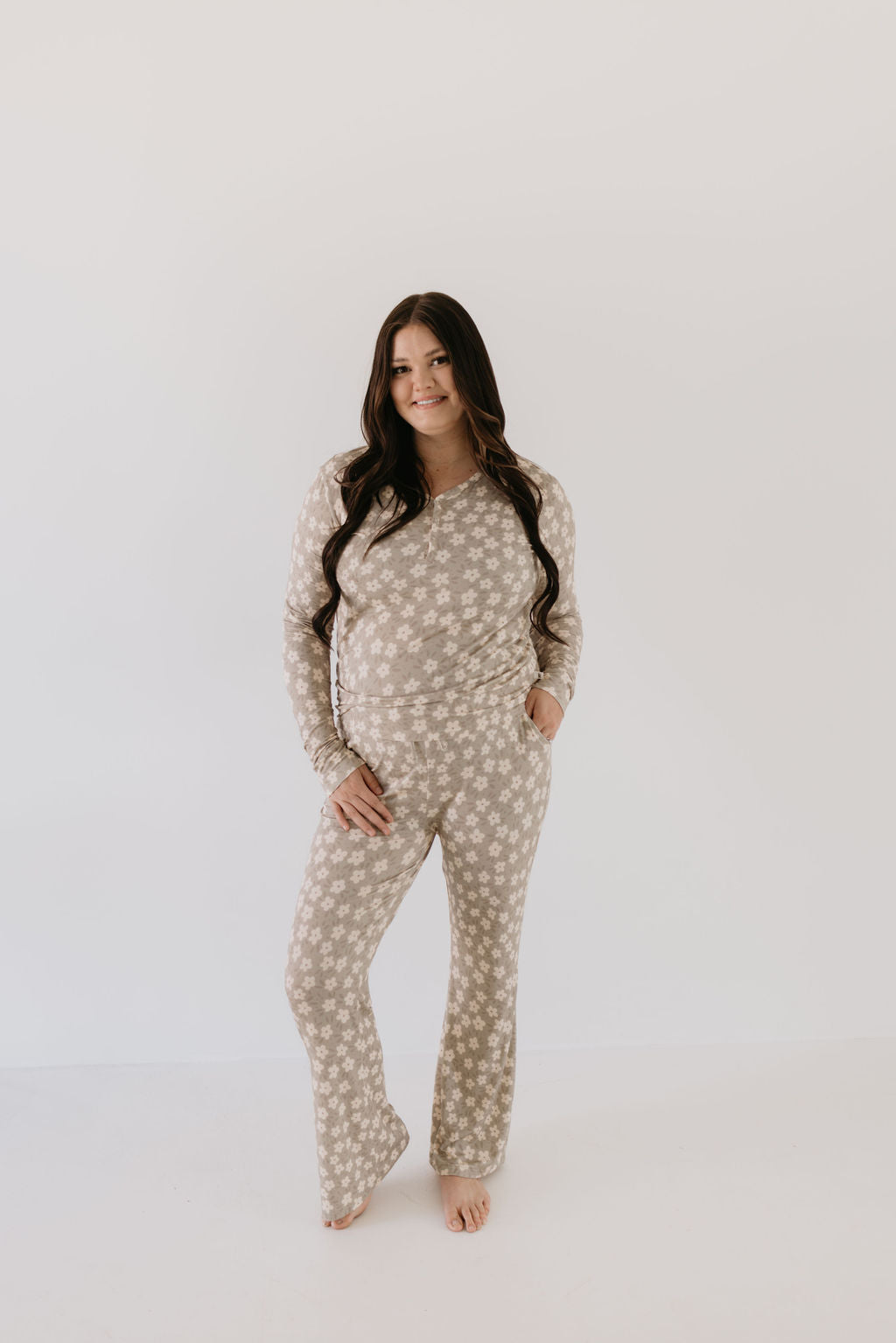 A woman with long brown hair stands barefoot against a white background, smiling and wearing forever french baby’s Bamboo Two Piece Pajamas in French Gray Floral, with one hand in her pocket to showcase the breathable sleepwear.