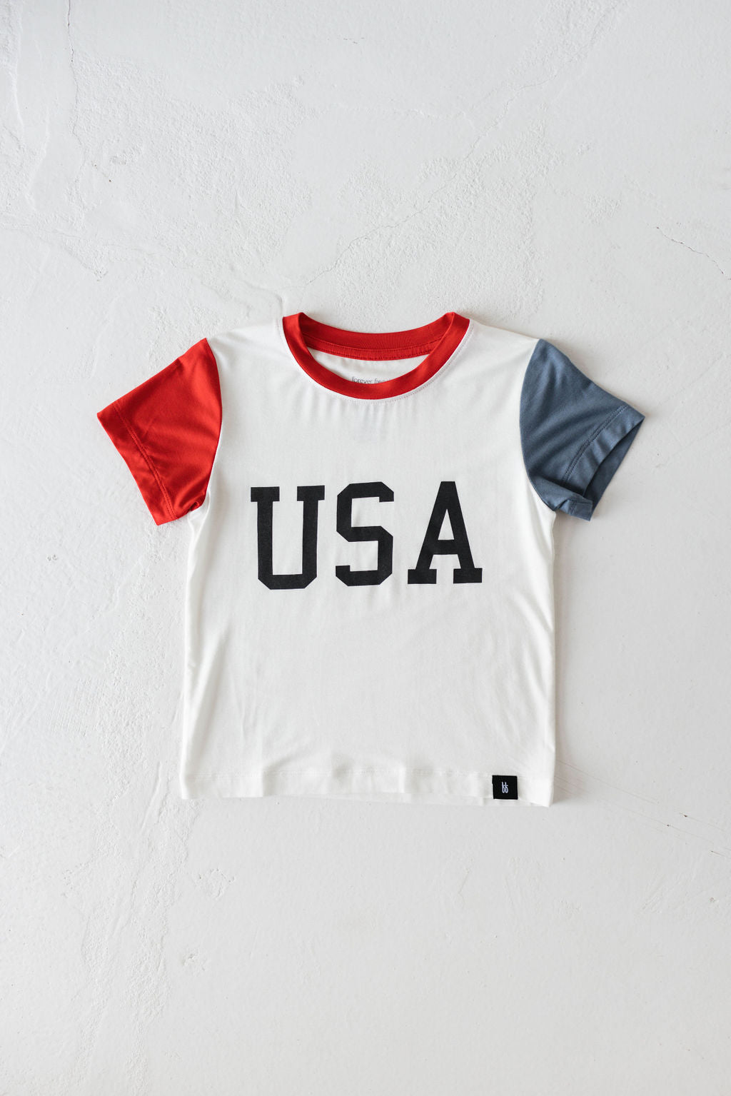 The Children's Bamboo T-Shirt | USA, Land That I Love by forever french baby is an eco-friendly white shirt designed for kids. It features the word "USA" boldly printed in black on the front, red short sleeves on one side, gray short sleeves on the other, and a matching red collar. Perfect for 4th of July celebrations, it is displayed against a plain white background.
