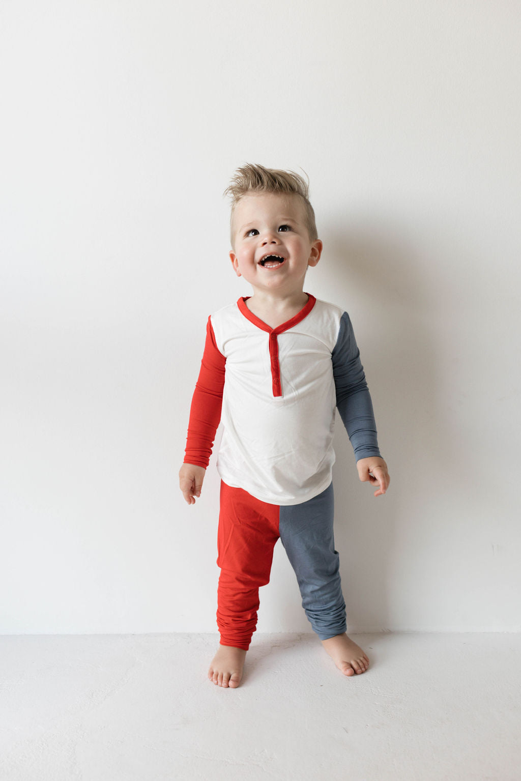 A smiling toddler stands against a white wall, wearing the "Bamboo Two Piece Pajamas | USA, Land That I Love" from forever french baby. The joyful child is dressed in a long-sleeved shirt with red sleeves and matching red and blue pants—perfect for 4th of July celebrations. The excitement and joy are evident as they look upward.