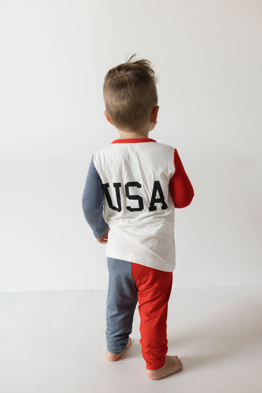 A young child stands facing away from the camera, wearing a white shirt with the letters "USA" printed on the back. The shirt has multicolored sleeves, one blue and one red, and the child is also wearing matching red and blue pants—perfect as festive 4th of July attire or as hypo-allergenic clothing for sensitive skin. They are sporting the Bamboo Two Piece Pajamas | USA, Land That I Love by forever french baby.