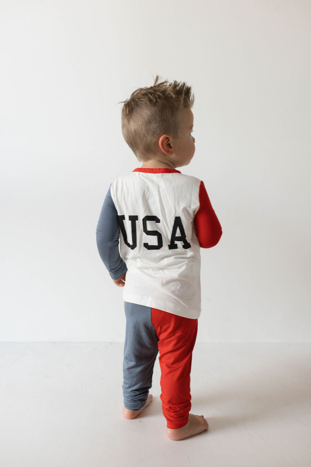 A young child stands on a white background, facing away. They are wearing 4th of July attire from forever french baby, featuring the Bamboo Two Piece Pajamas called "USA, Land That I Love." The white shirt has "USA" printed in black on the back, with one sleeve blue and the other red. The hypo-allergenic clothing includes two-toned pants with one leg gray and the other red.