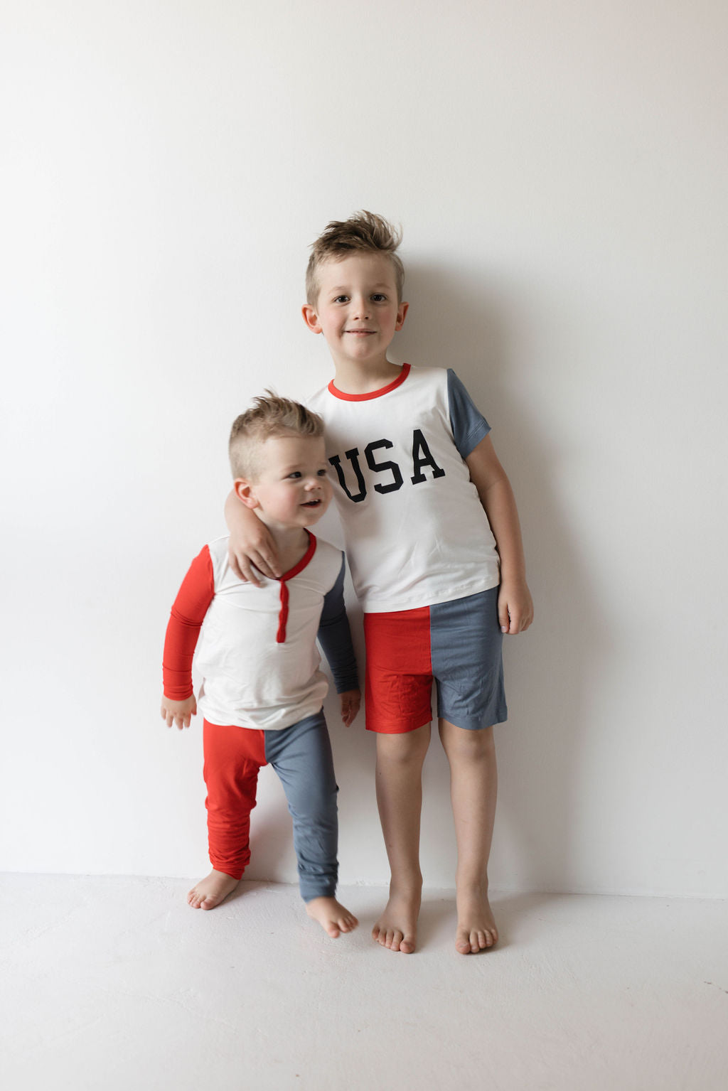 Two young children stand against a white wall. The older child is wearing the "Bamboo Short Set | USA, Land That I Love" by forever french baby, featuring a white shirt with "USA" printed on it and half red, half blue shorts. The younger child is dressed in mismatched red and blue kids pajamas. The older child has an arm around the younger child's shoulder.