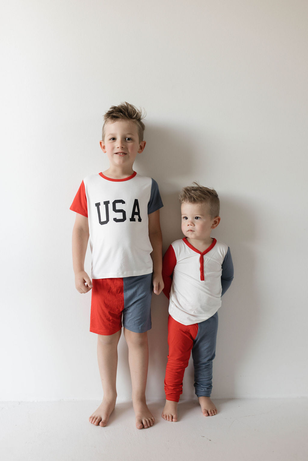 Two young boys stand against a white wall. The older boy wears the "USA, Land That I Love" Bamboo Two Piece Pajamas from forever french baby, featuring a white top with "USA" printed on it and matching red, white, and blue shorts—ideal for 4th of July celebrations. The younger boy also dons hypo-allergenic clothing: a white long-sleeve top paired with red and blue pants. Both have short, styled blonde hair.