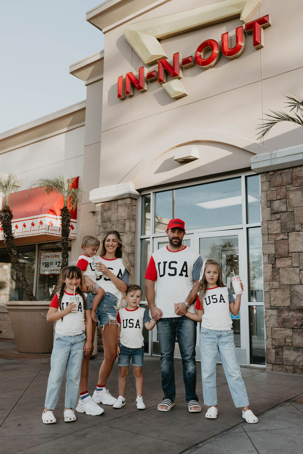 A family of seven, each donning festive 4th of July attire and casual wear, stands in front of an In-N-Out Burger restaurant. The parents are holding two young children, who are dressed in the "Children's Bamboo T-Shirt | USA, Land That I Love" by forever french baby. Meanwhile, the three older children stand beside them, each clutching a beverage cup with the restaurant's signage prominently displayed above.