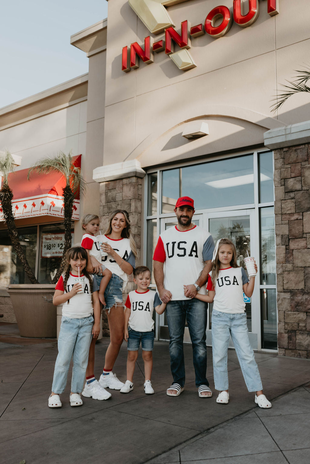 A family of six stands in front of an In-N-Out Burger restaurant, all wearing matching USA-themed outfits: white "Children's Bamboo T-Shirts | USA, Land That I Love" from forever french baby and blue jeans. Their eco-friendly shirts give a nod to sustainability. They hold hands and look at the camera, smiling with the In-N-Out logo behind them.