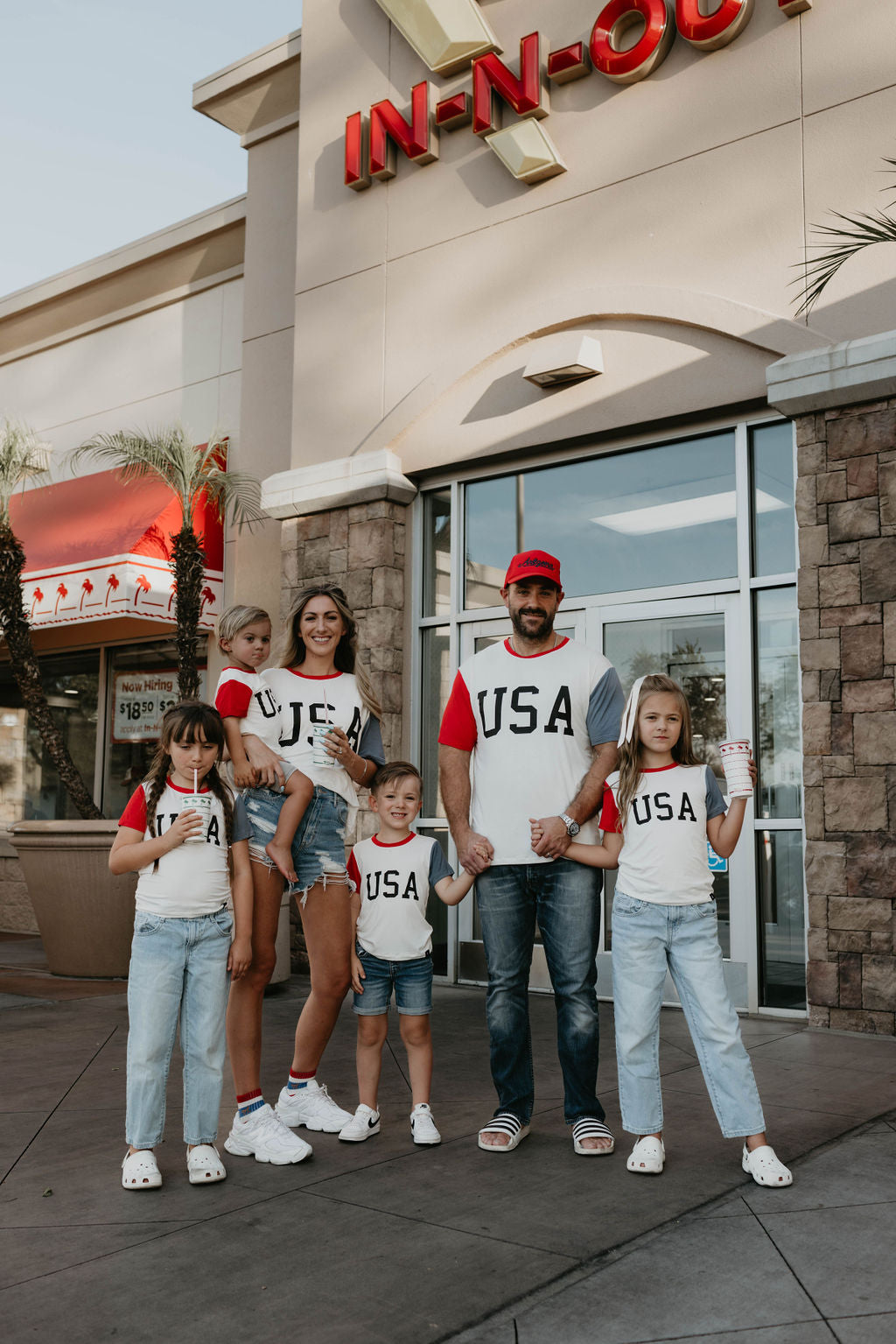 A family of six, two adults and four children, stand outside an In-N-Out Burger restaurant. They are all wearing matching Adult Bamboo T-Shirts with the stylish USA "Land That I Love" design on the front from the brand forever french baby. The children are holding drinks, and one child is proudly clutching a burger.