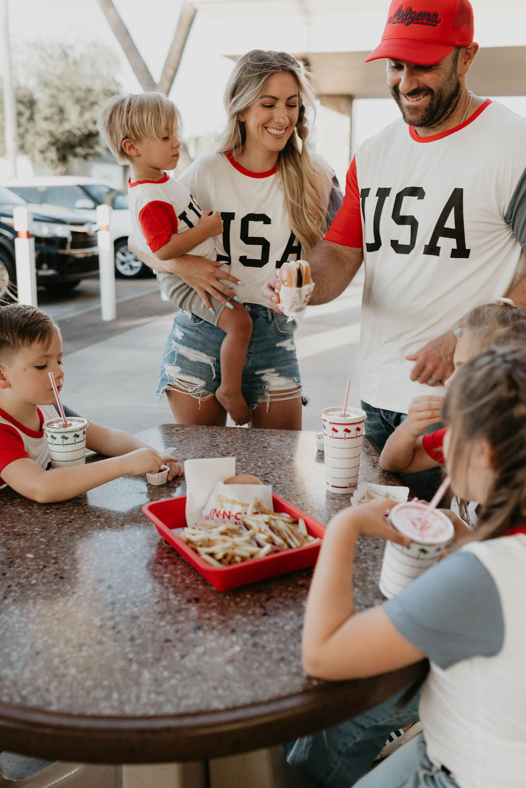 A smiling family of five enjoys a meal outdoors at a round table. The parents and their three young children, all wearing matching "Children's Bamboo T-Shirts | USA, Land That I Love" from forever french baby, are gathered around with drinks and fries. The mother holds a toddler while the father holds a burger.