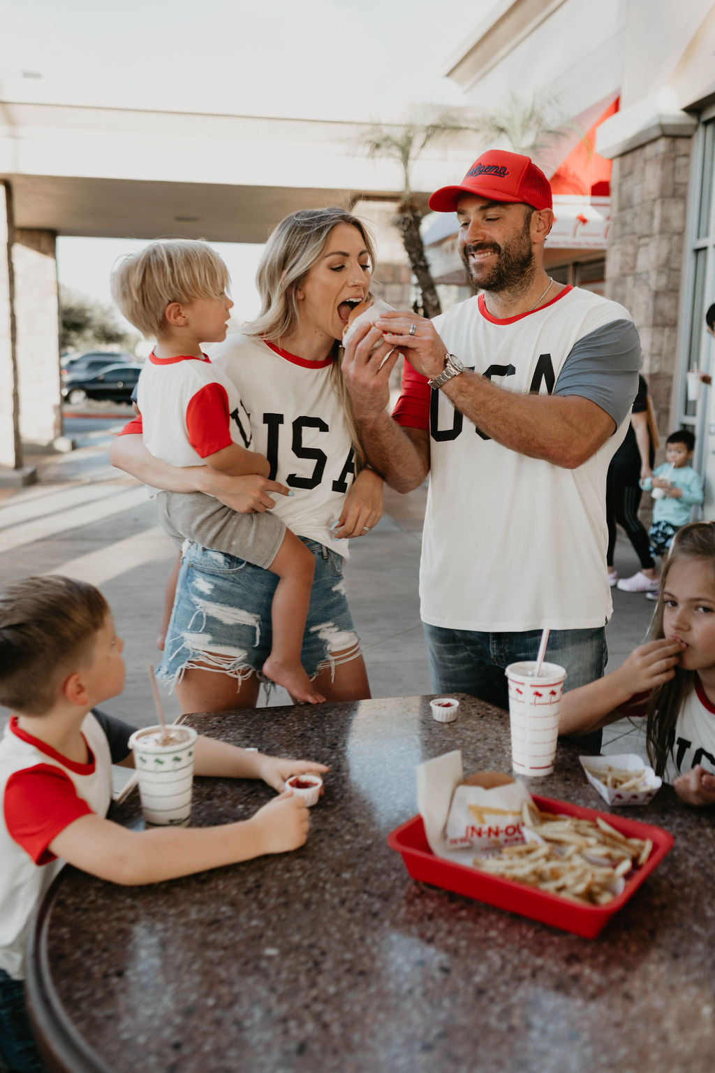 A family of five, wearing matching "Children's Bamboo T-Shirt | USA, Land That I Love" from forever french baby, enjoys a meal at an outdoor table of a fast-food restaurant. The parents stand while the mother eats a fry offered by the father. Two young boys and a girl sit at the table with food and drinks, their eco-friendly shirts adding to their festive spirit.