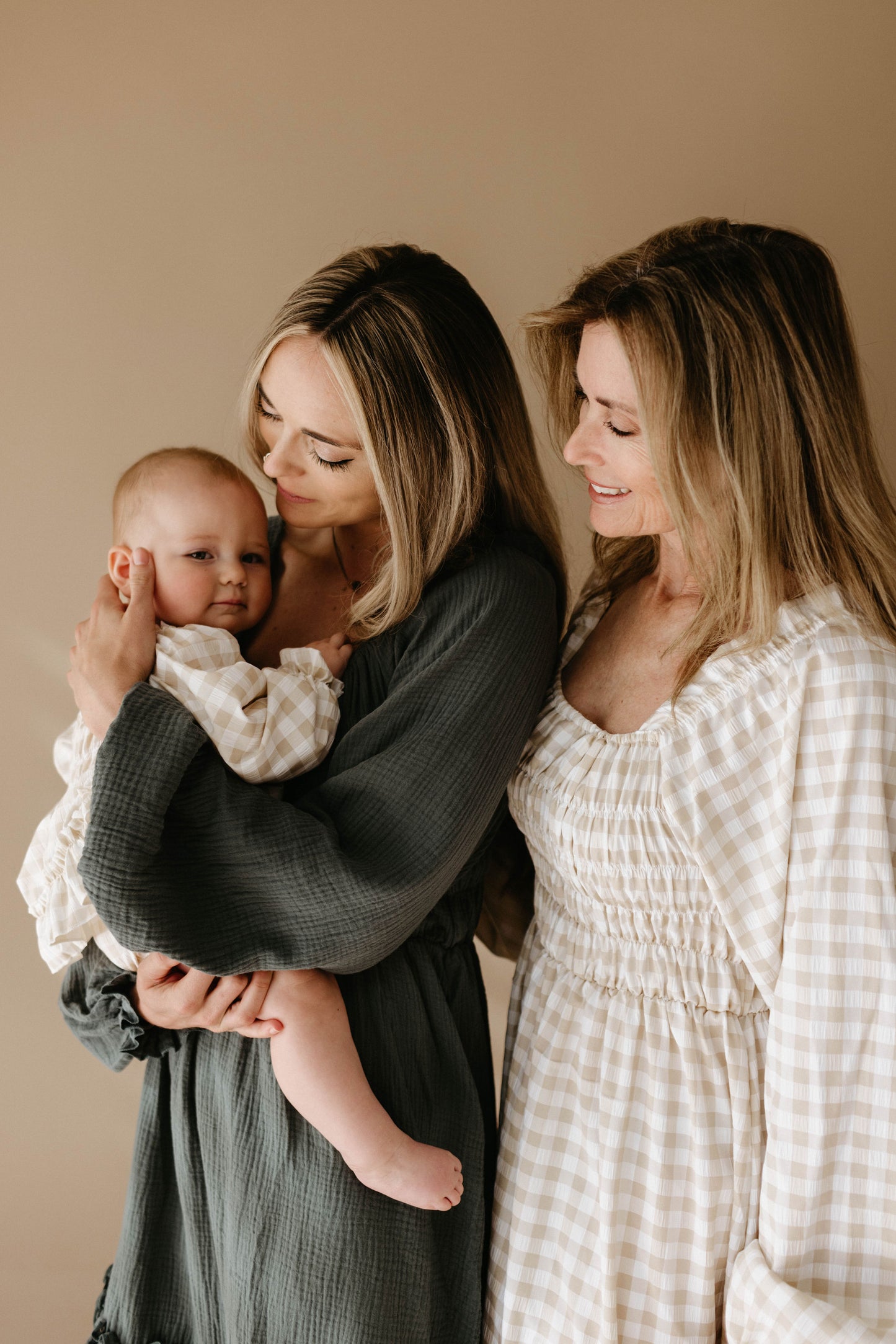 Two women with long, blonde hair smile warmly at a baby, one holding the baby in her arms. Both wear light, neutral-toned Women's Dress | Meadow by forever french baby against a plain tan background.