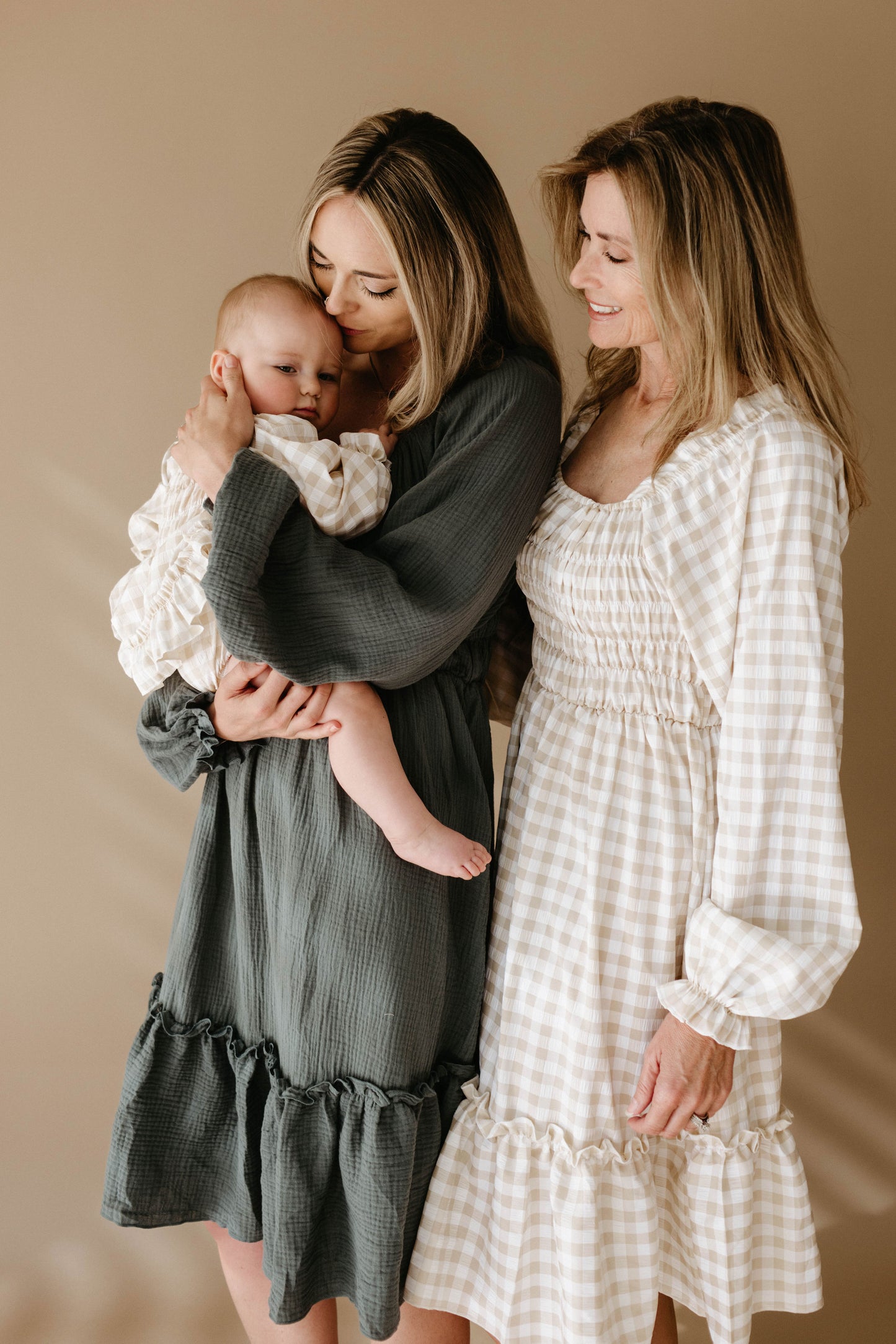 Two women with long blonde hair, dressed in light-colored Meadow dresses from forever french baby, smile warmly at each other while one holds an infant. They stand against a plain beige background.