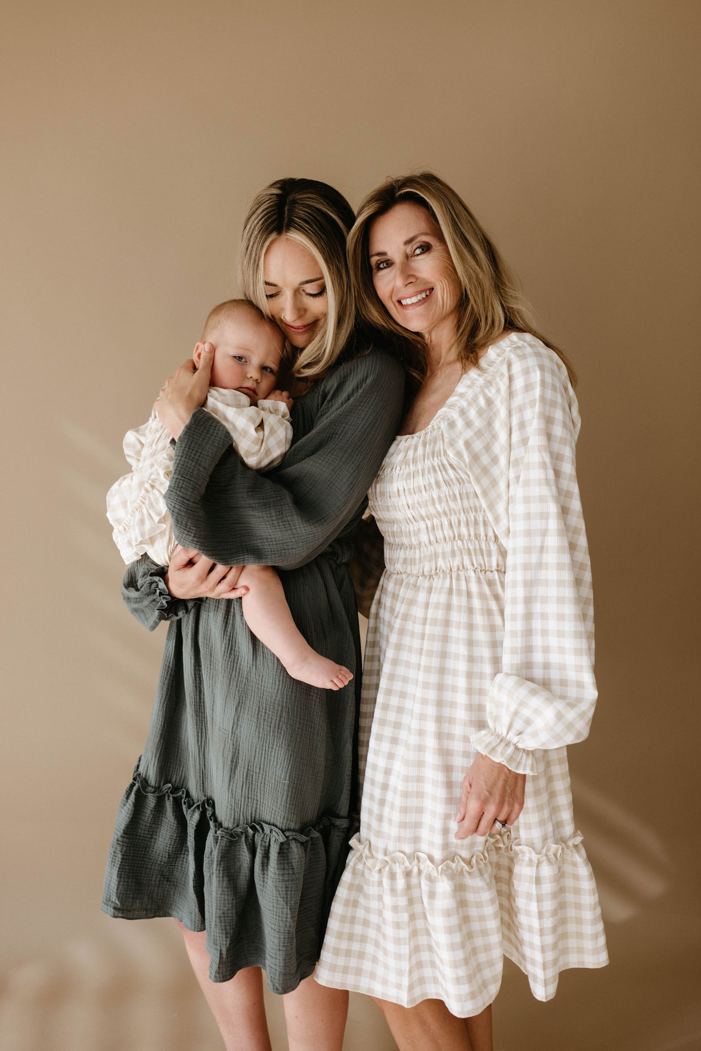 Three generations of women in Meadow long-sleeved ruffled dresses by forever french baby stand together—one holding a baby—all dressed in neutral tones against a simple beige background.