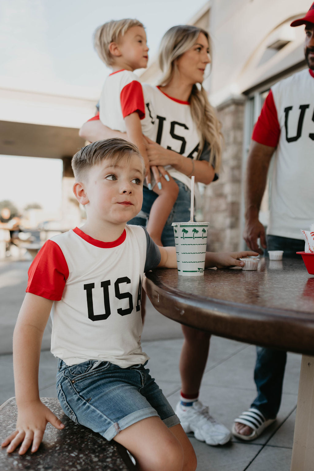 A young boy in a "Children's Bamboo T-Shirt | USA, Land That I Love" by forever french baby and denim shorts sits at a round outdoor table holding a drink cup. Behind him, a woman holds a toddler, also in matching 4th of July clothes from the same brand, standing next to a man with a beard. They are gathered in a casual, sunny setting.