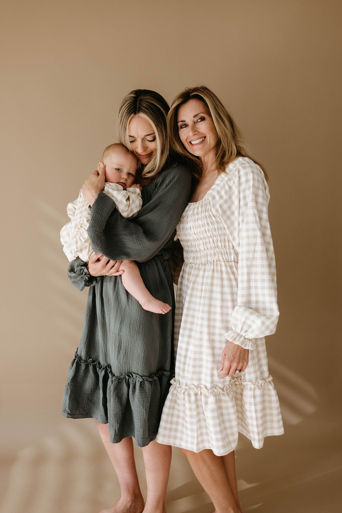Three generations of women pose barefoot against a beige background: a smiling older woman in a white gingham dress, and a younger woman in the forever french baby Women's Dress | Meadow holding a baby.