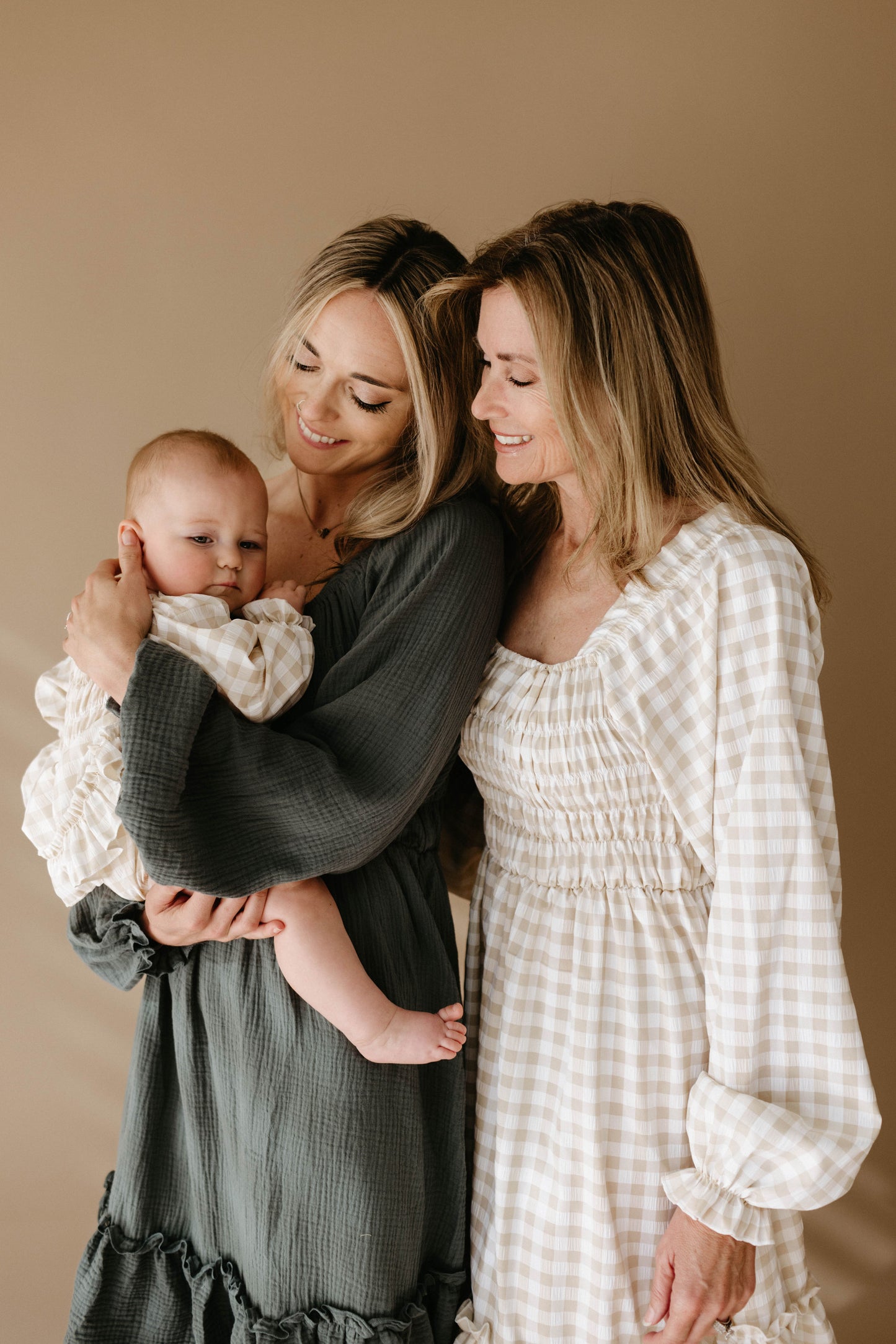 A smiling woman holding a baby stands beside an older woman, all dressed in forever french baby “Women's Dress | Meadow” long-sleeve dresses against a beige background, radiating warmth and affection.