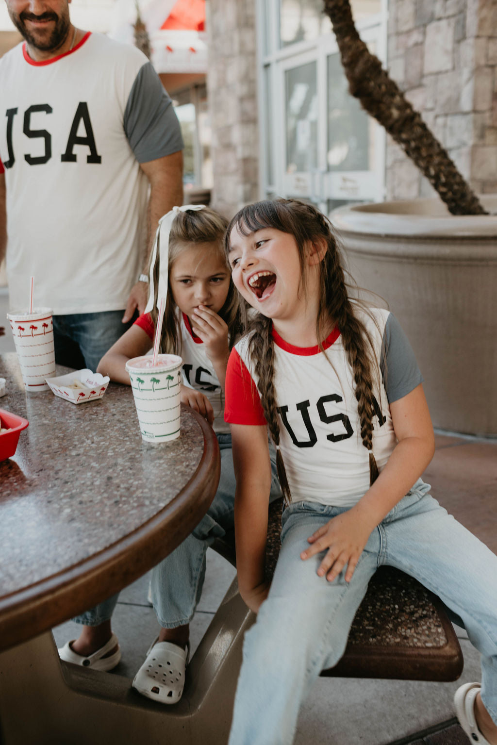 Two young girls in matching "USA, Land That I Love" Children's Bamboo T-Shirts from forever french baby sit at an outdoor table with drinks and snack containers. One laughs heartily, while the other looks on curiously. An adult in a similar eco-friendly shirt made from bamboo stands nearby, with an arm resting on the table.