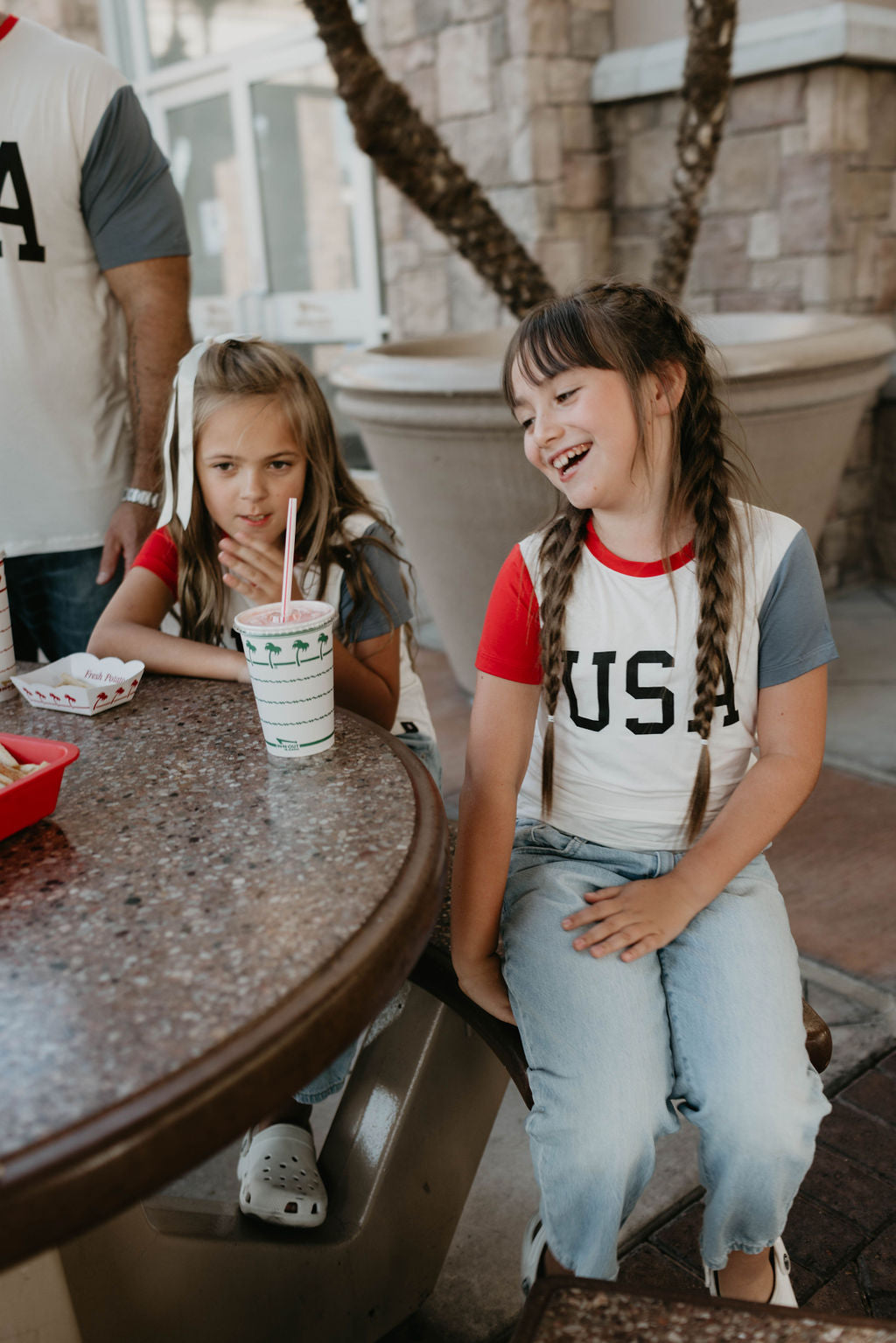 Two girls with braids sit at a round table outdoors. The girl on the left looks at a drink while the girl on the right, wearing a forever french baby Children's Bamboo T-Shirt with "USA, Land That I Love" on it, laughs. Part of an adult and a flowerpot are visible in the background.