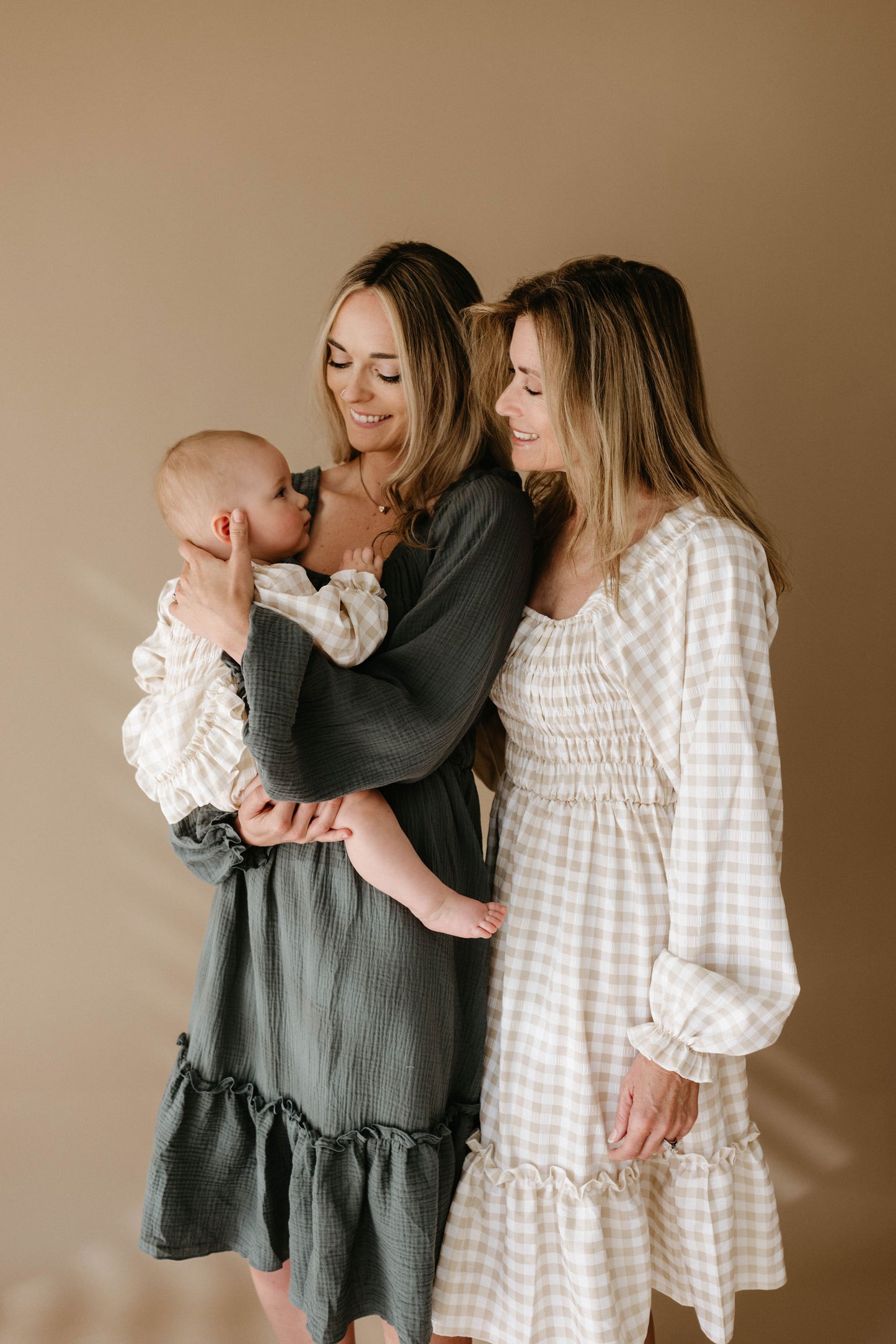 Two women smile against a beige backdrop. The woman on the left wears the Meadow dress by forever french baby, while the woman on the right is in a light checked dress. One holds a baby dressed in cream.