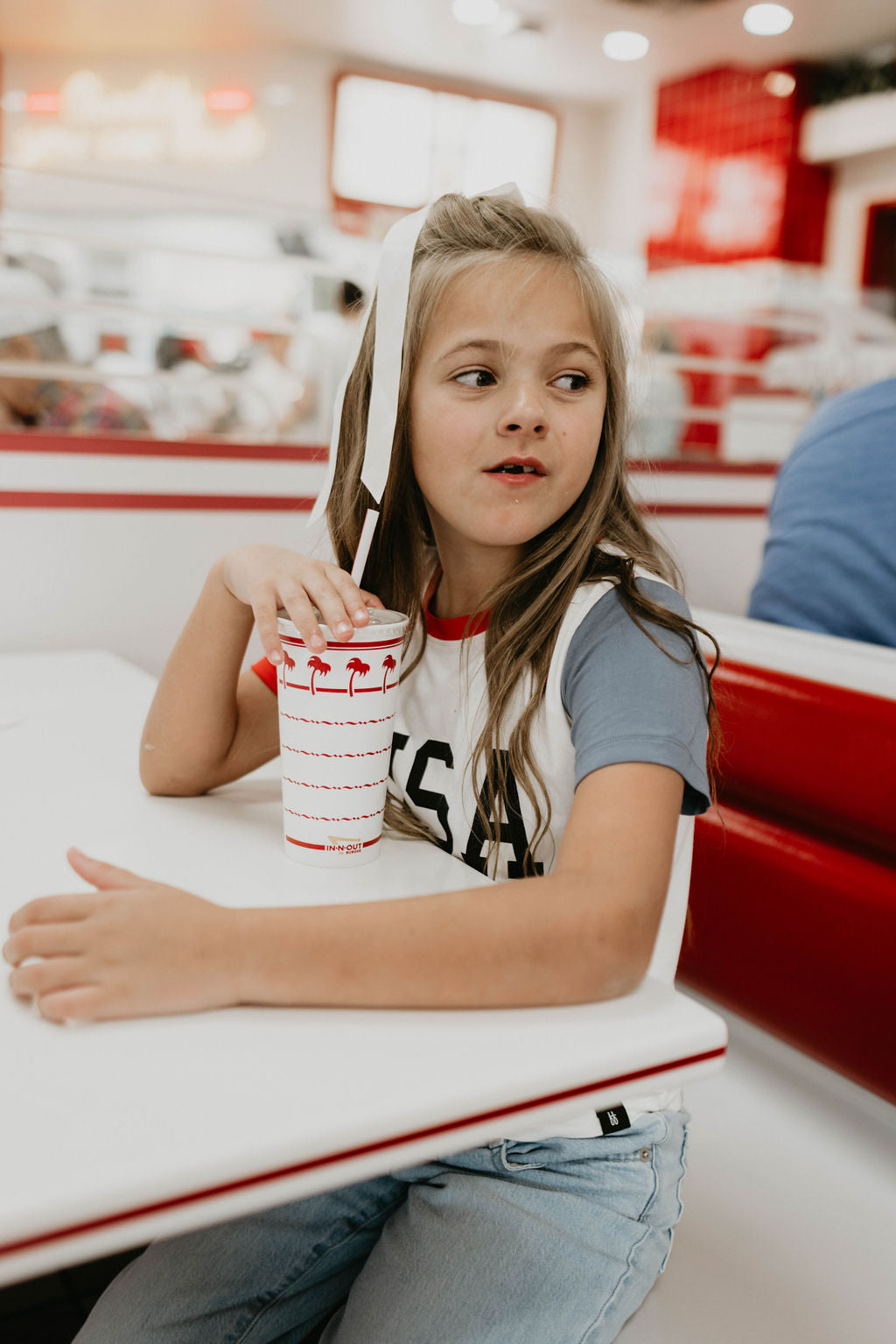 A young girl with long brown hair, wearing a white and blue Children's Bamboo T-Shirt from forever french baby, sits at a table in a fast-food restaurant. She holds a cup with a red straw and palm tree design, looking to her left with her mouth slightly open while resting her arm on the table.
