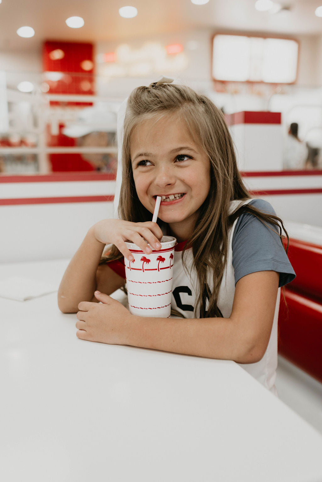 A young girl with long hair sits at a table in a brightly-lit restaurant, smiling while drinking from a cup with a straw. Dressed in an eco-friendly Children's Bamboo T-Shirt by forever french baby featuring the "USA, Land That I Love" design, she enjoys the festive red and white decor, surrounded by people and lights.