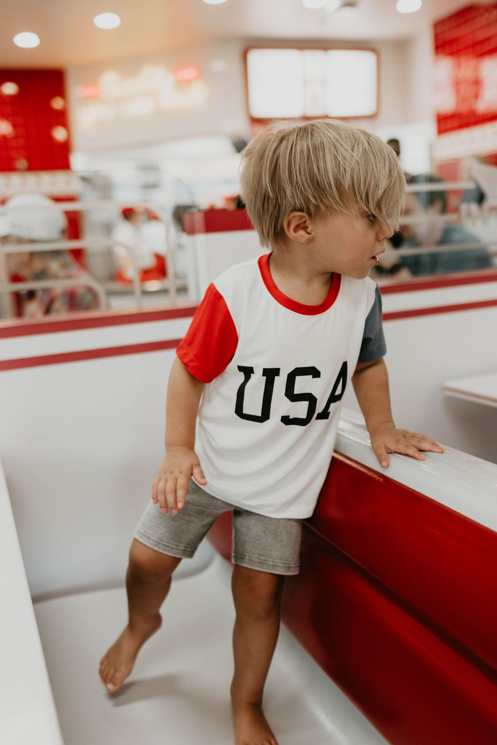 A young child with blonde hair, wearing an eco-friendly Children’s Bamboo T-Shirt by forever french baby with "USA, Land That I Love" printed on the front and gray shorts, stands on the seating area of a bright and modern diner or restaurant. The background shows people dining amidst vibrant red and white decor.