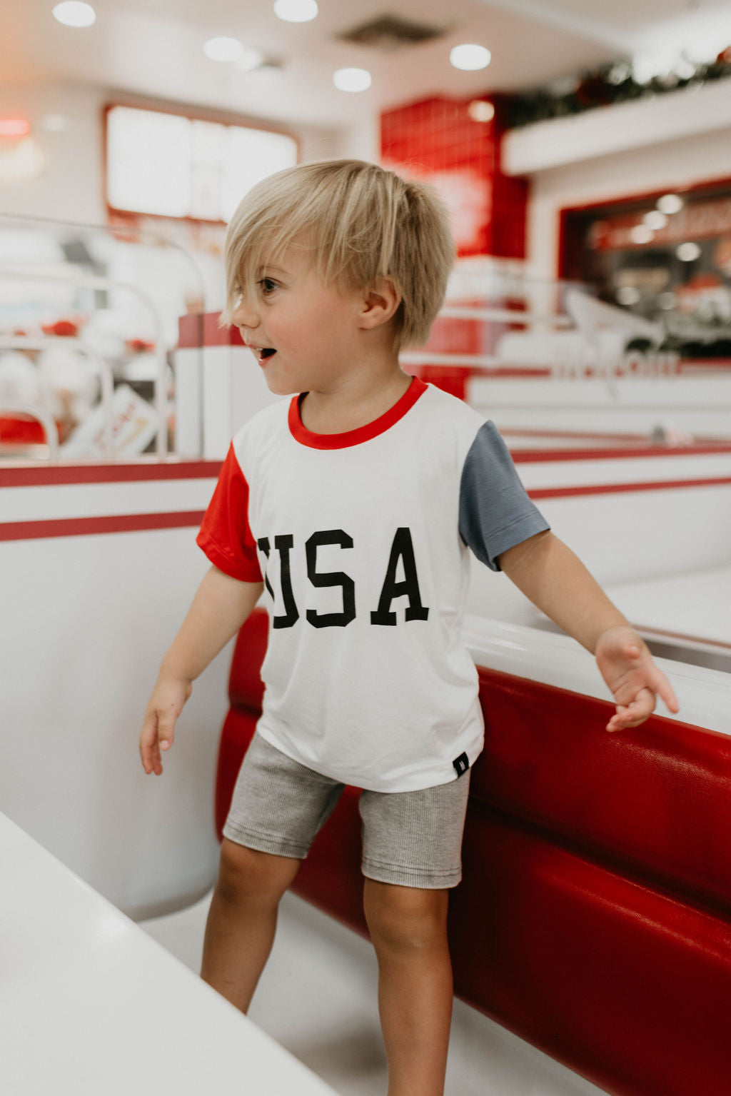 A young child with blonde hair stands inside a diner setting, wearing a forever french baby Children's Bamboo T-Shirt | USA, Land That I Love with red and blue sleeves and grey shorts. The decor of the diner is primarily red and white, perfectly complementing their 4th of July clothes.