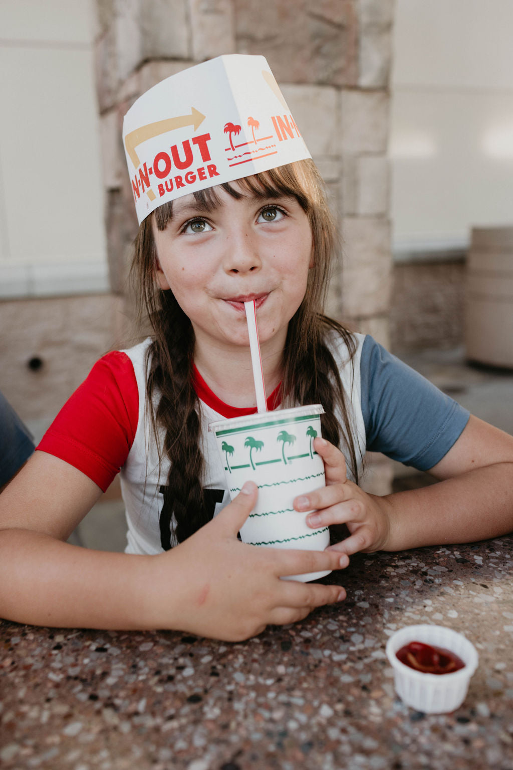 A young girl with long brown hair, wearing an In-N-Out Burger paper hat and a colorful Children's Bamboo T-Shirt by forever french baby, featuring the design "USA, Land That I Love," sits at a table sipping from a cup with a straw. A small container of ketchup is on the table next to her.