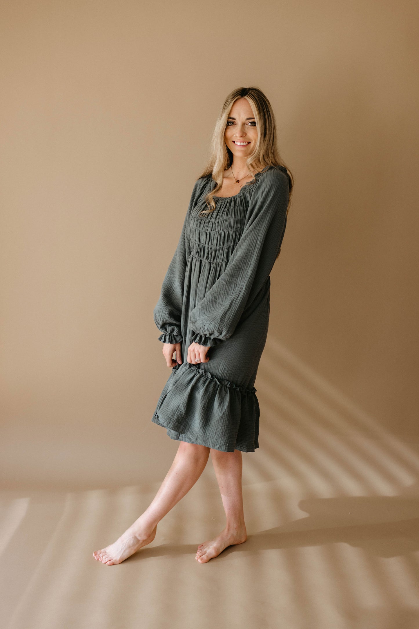 A woman with long blonde hair stands barefoot on a beige floor, wearing the Women's Dress | Meadow by forever french baby. She smiles gently, posing with one foot slightly raised against a softly lit beige background.