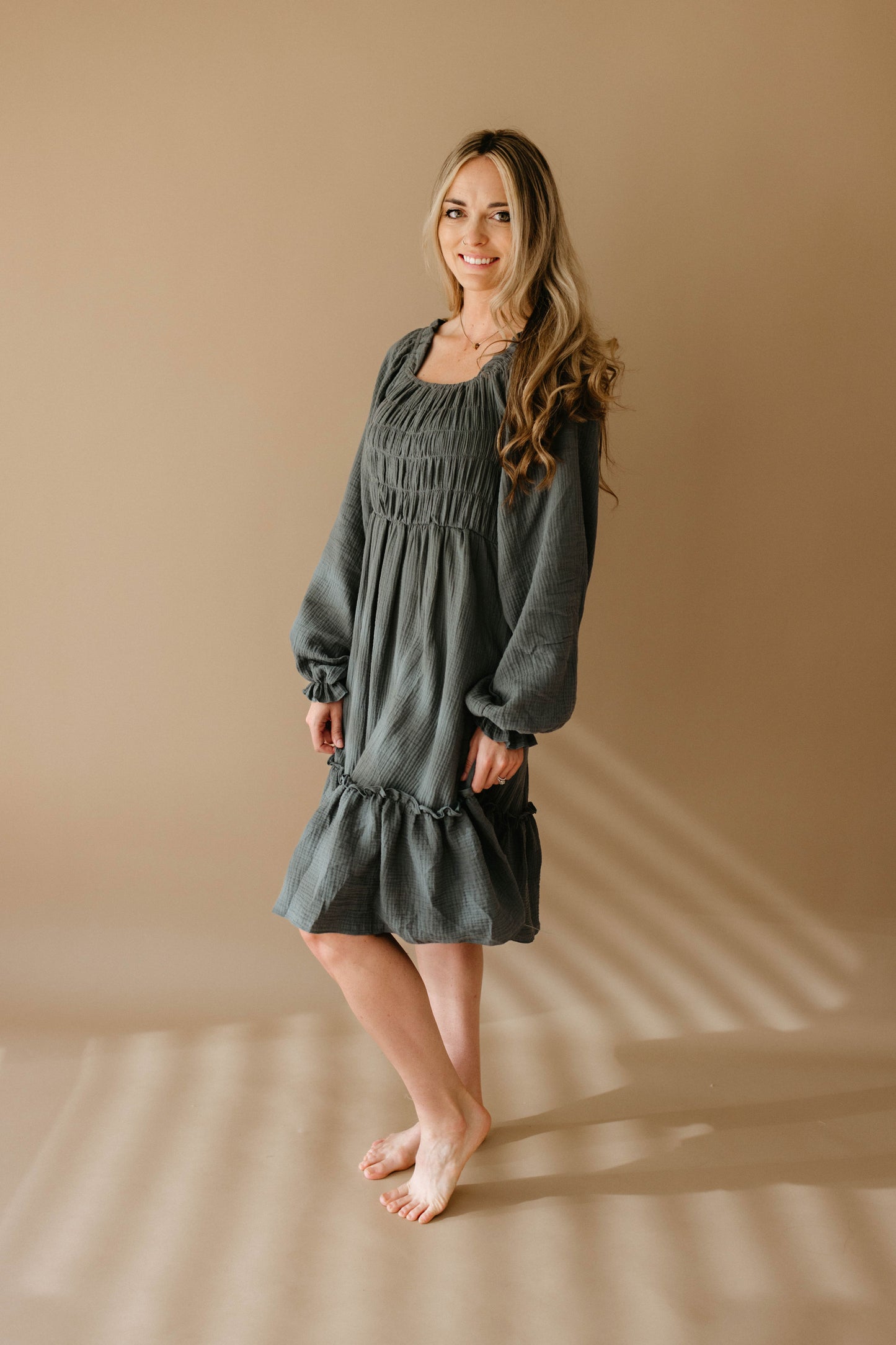 A woman with long wavy hair stands barefoot on a beige floor, wearing the forever french baby Women's Dress | Meadow in dark gray. She smiles slightly against a plain beige background.