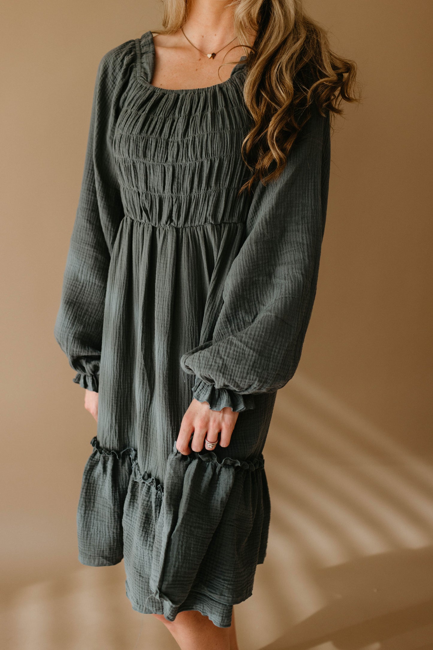 A woman models the forever french baby Women's Dress | Meadow, a long-sleeved dark green smocked dress, standing against a beige background with her face partially out of frame as she gently holds the skirt with one hand.