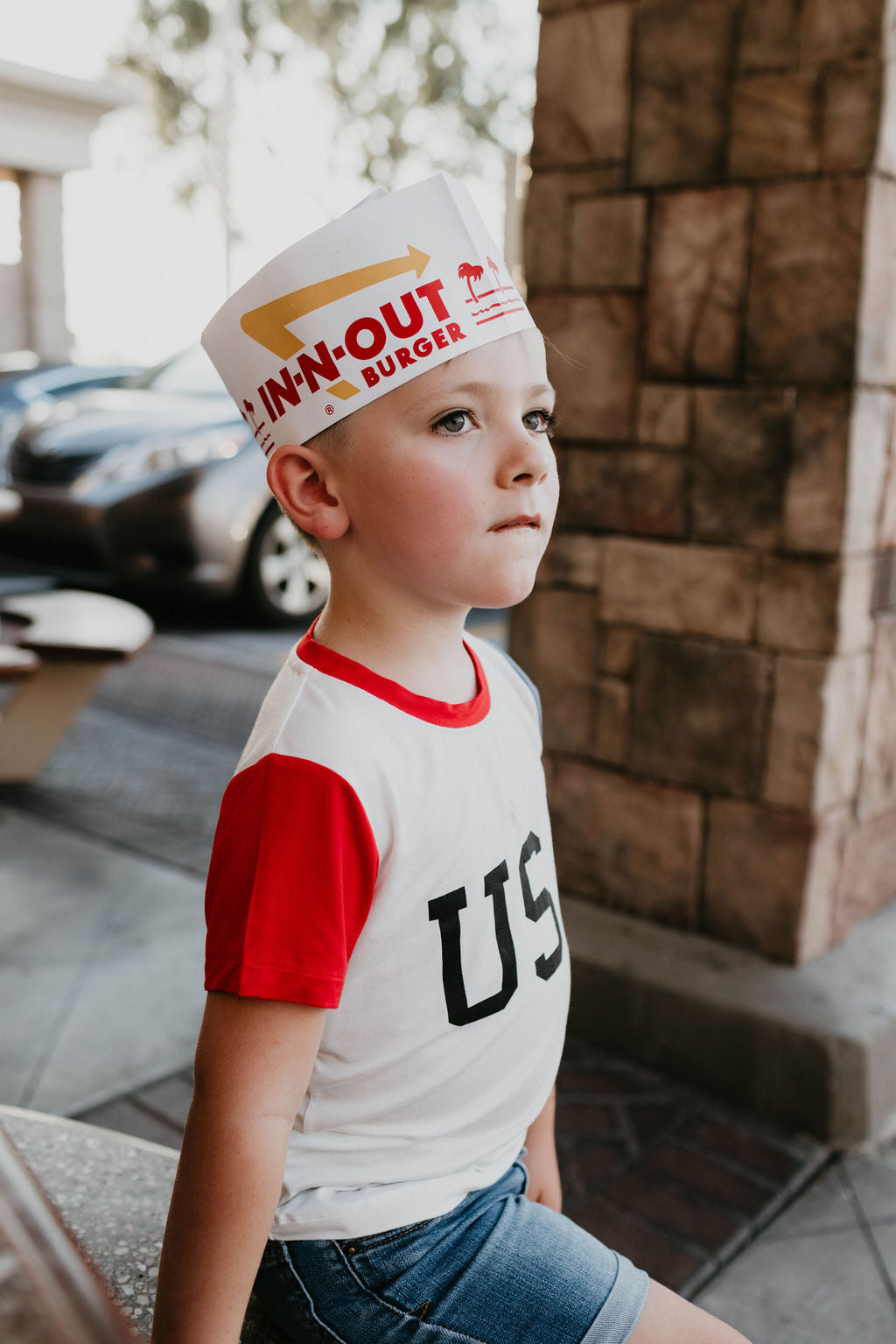 A young boy sits outside wearing an In-N-Out Burger paper hat. He is dressed in a forever french baby Children's Bamboo T-Shirt with red sleeves featuring the letters "USA" on the front and denim shorts, perfect 4th of July clothes. The background shows a stone pillar and a parked car.
