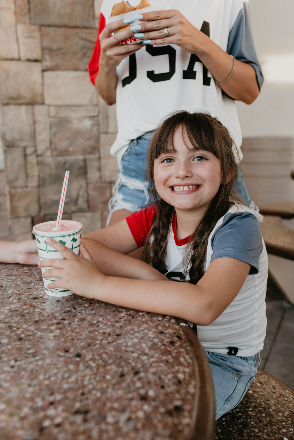 A smiling young girl with braided hair sits at a table, holding a cup with a straw. She is wearing the Children's Bamboo T-Shirt by forever french baby, which has red and blue sleeves. Another person, wearing the "USA, Land That I Love" shirt from the same brand, stands behind her holding a sandwich. They are outdoors near a stone wall.