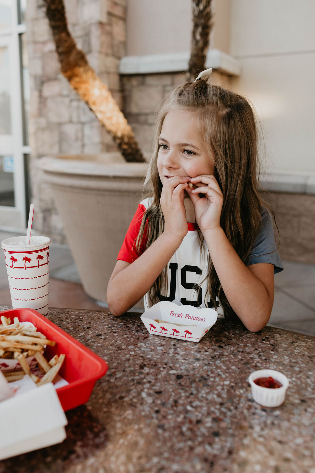 A young girl with long hair sits at a table outdoors, enjoying a meal. She is eating fries from a red tray with a cup of soda beside her. She wears the **Children's Bamboo T-Shirt | USA, Land That I Love** from **forever french baby**, an eco-friendly shirt perfect for 4th of July celebrations. The table has a small ketchup container nearby.