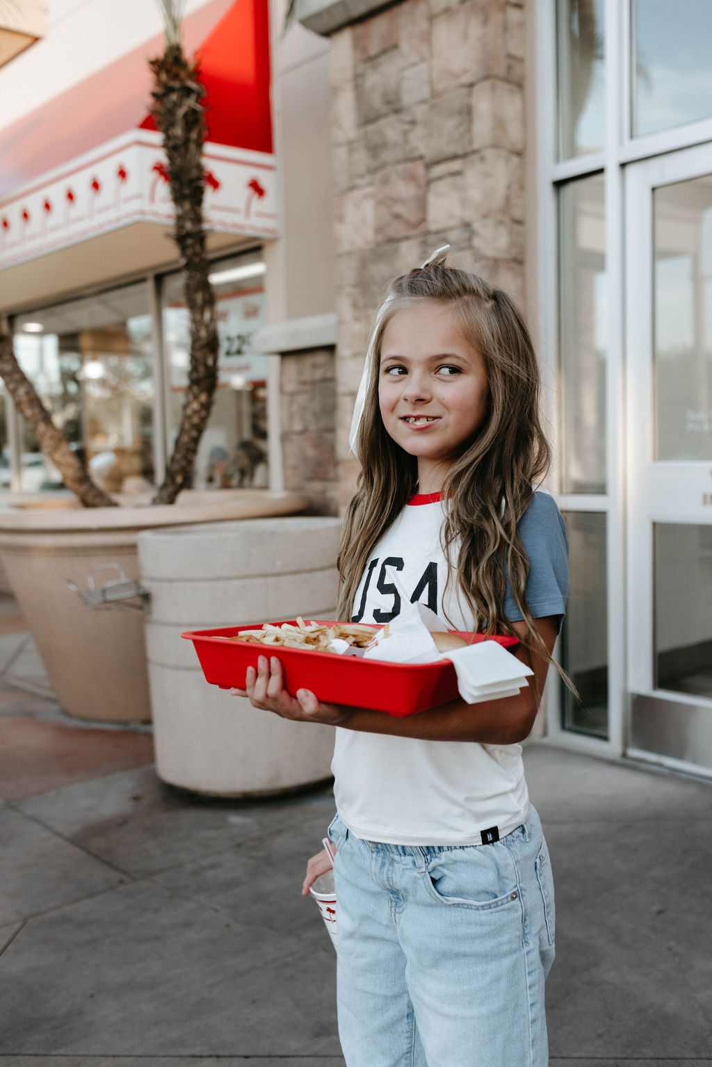 A young girl with long hair stands outside a building holding a red tray with food. She is wearing the forever french baby's Children's Bamboo T-Shirt featuring "USA, Land That I Love" and light blue jeans. Behind her are large potted plants and a glass window, suggesting a casual, outdoor dining area.